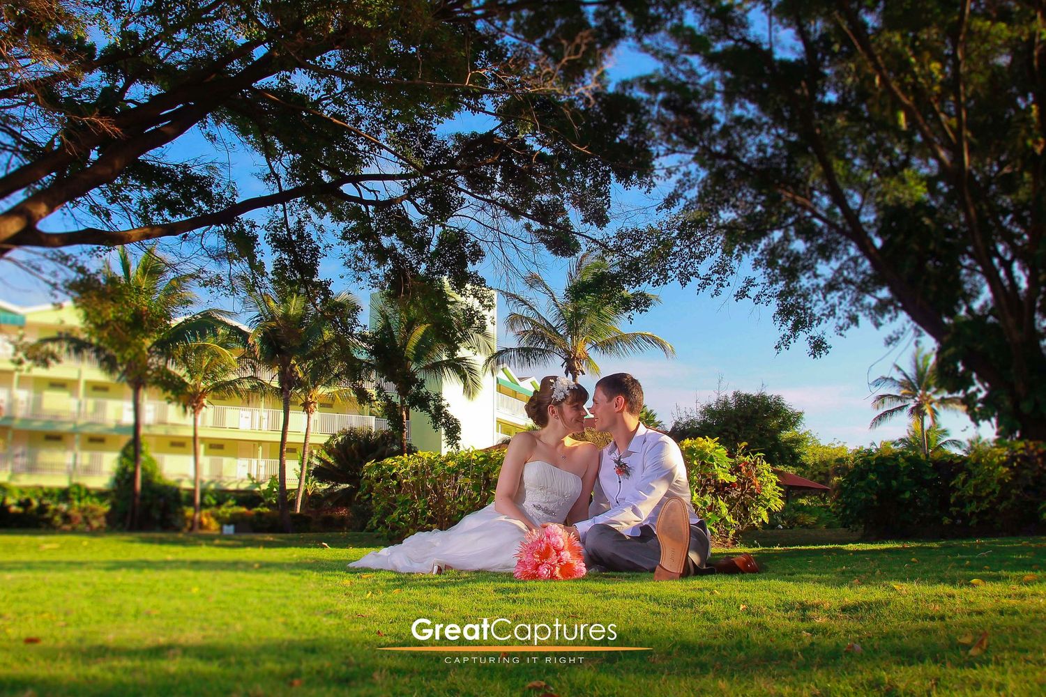 Bride and groom sitting on lush green grass under tropical trees at a Hawaiian resort wedding photo by Great Captures.