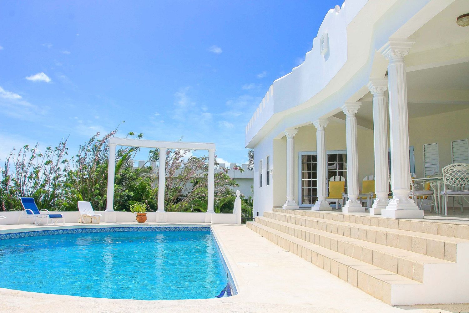 Luxury white villa with blue swimming pool, elegant columns, and tropical landscaping under a bright blue sky.