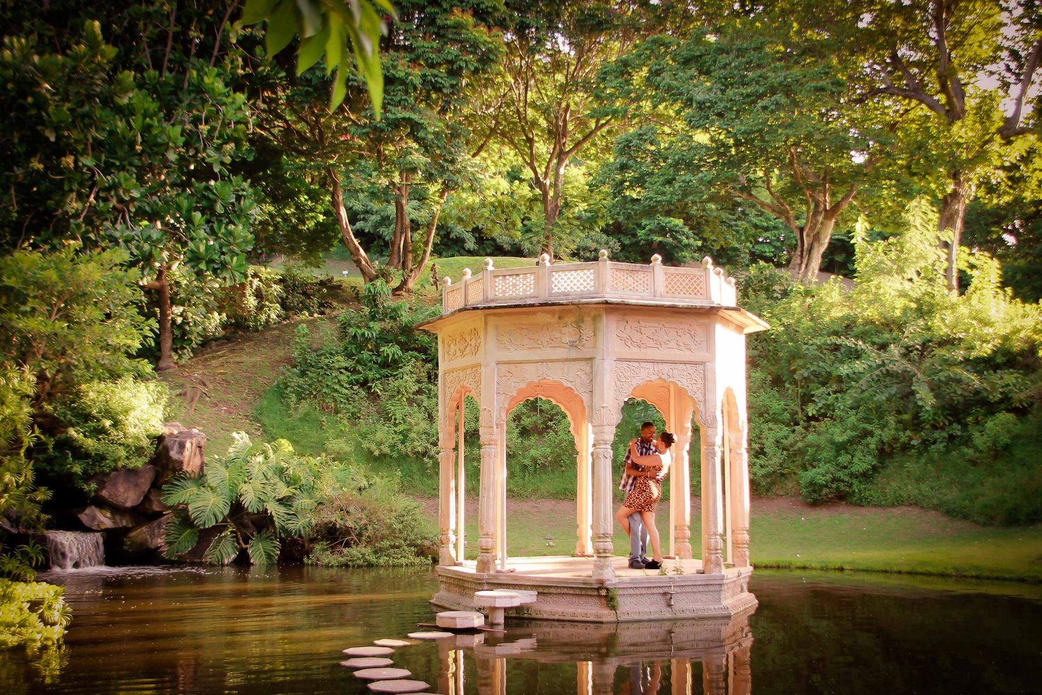 Couple embracing in an ornate white gazebo on a tranquil pond surrounded by lush green trees.