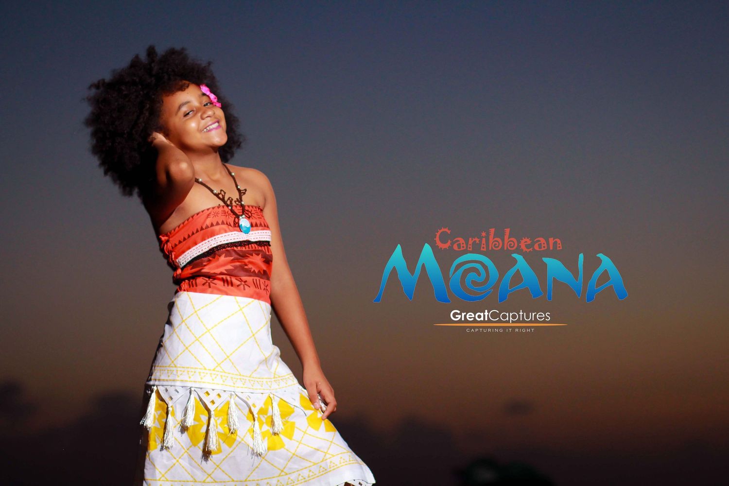 Young girl dressed as Caribbean Moana in colorful costume, smiling against a sunset sky backdrop.