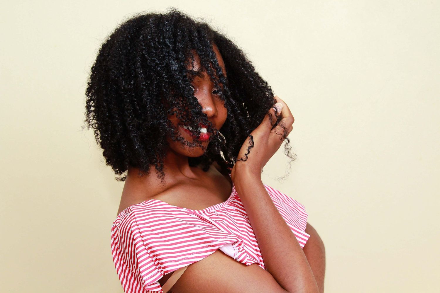 Young Black woman with curly natural hair in red striped off-shoulder top posing against beige background.