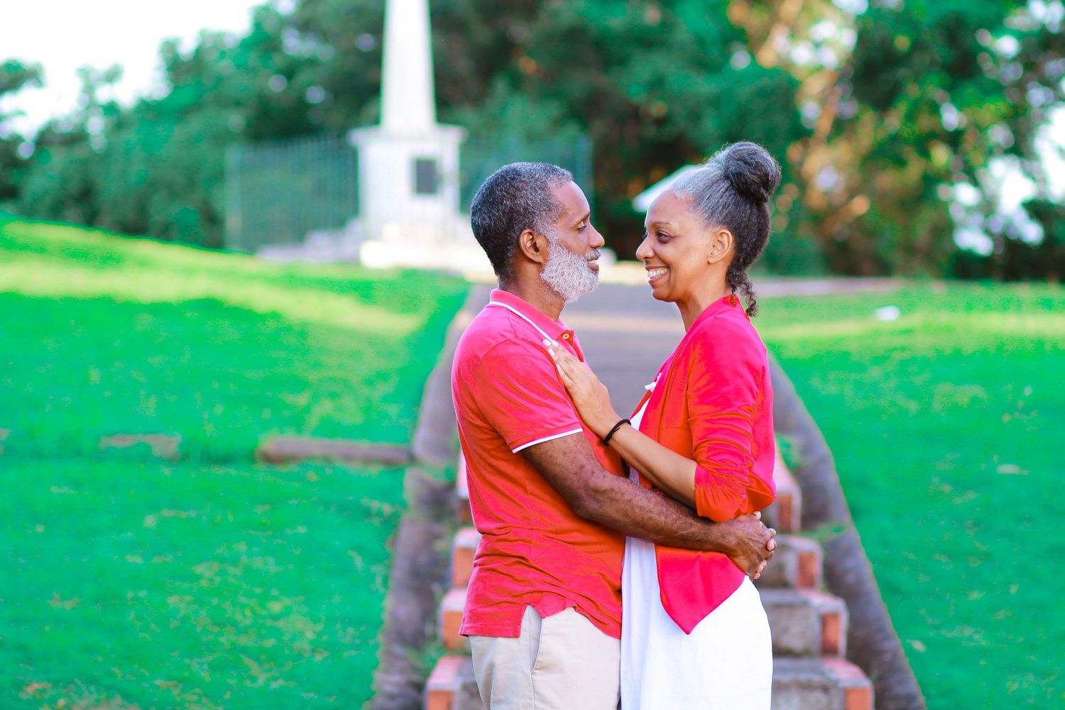Elderly couple in matching red shirts embracing outdoors in a lush green park near a historic monument.