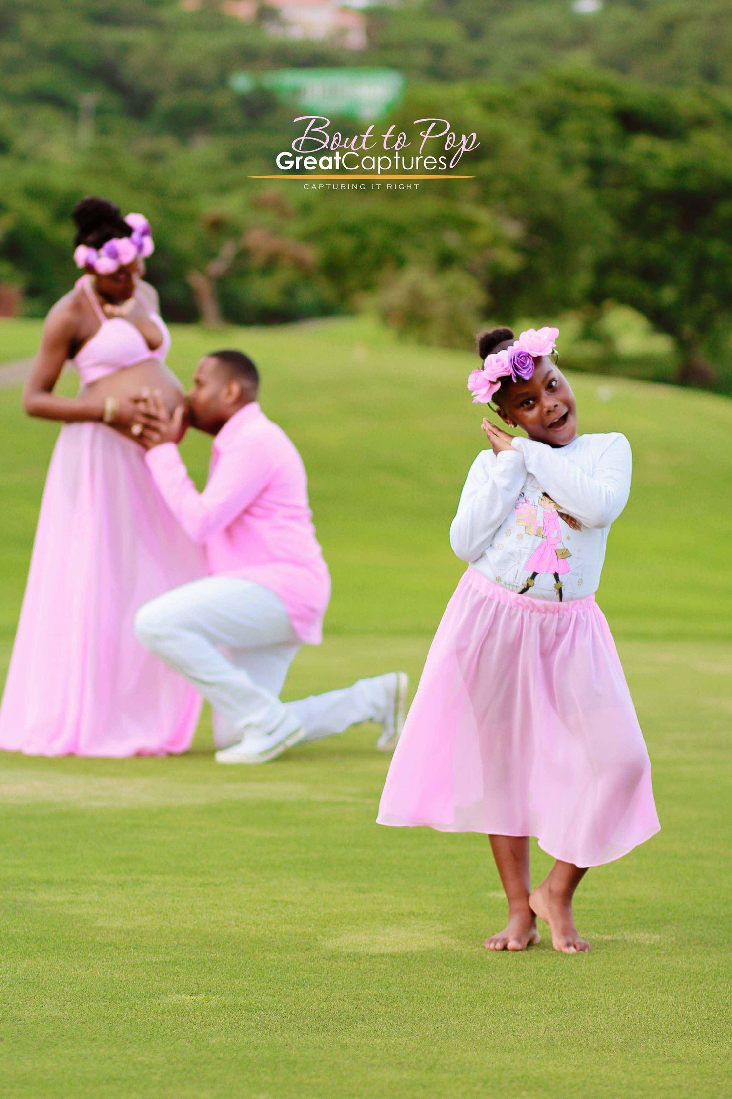 Family maternity photoshoot in pink outfits on golf course with adorable daughter in foreground.