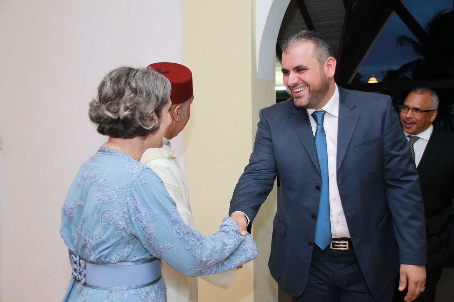 Man in suit shaking hands with woman in blue dress at formal diplomatic reception event.