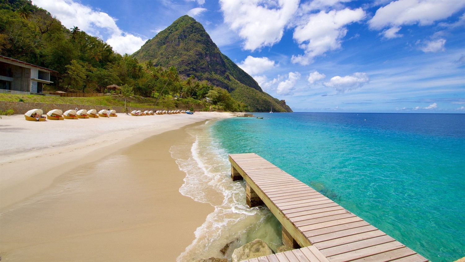 Pristine white sand beach with turquoise waters, wooden pier, and lush volcanic mountain in tropical St. Lucia.