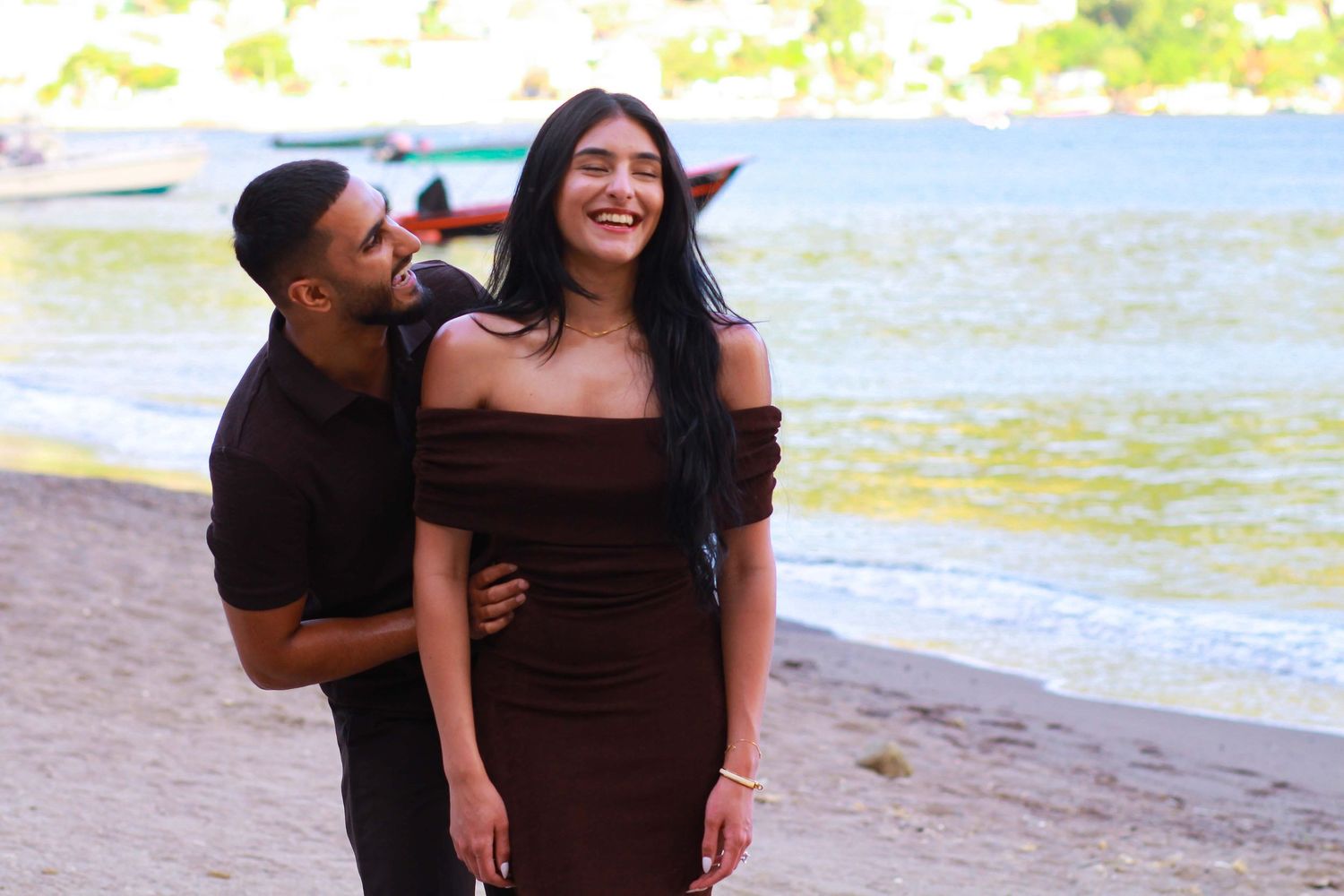 Couple posing on a sandy beach, woman in brown off-shoulder dress, man in black shirt, with water in background.