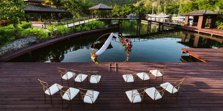 Elegant outdoor wedding ceremony setup on wooden deck overlooking serene pond with white chairs and floral arch.