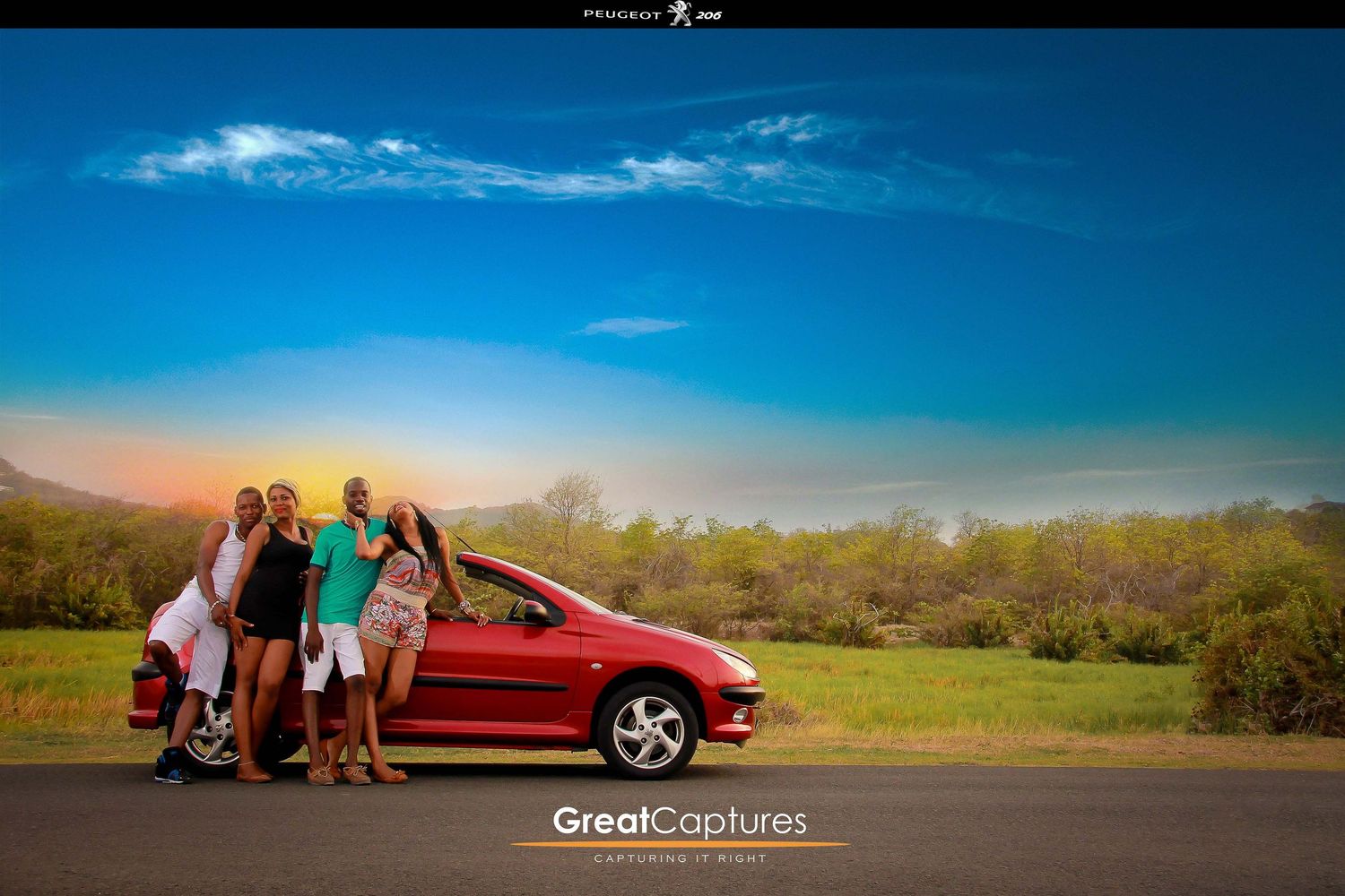 Four friends pose beside a red car on a scenic road with lush greenery and vibrant blue sky at sunset.
