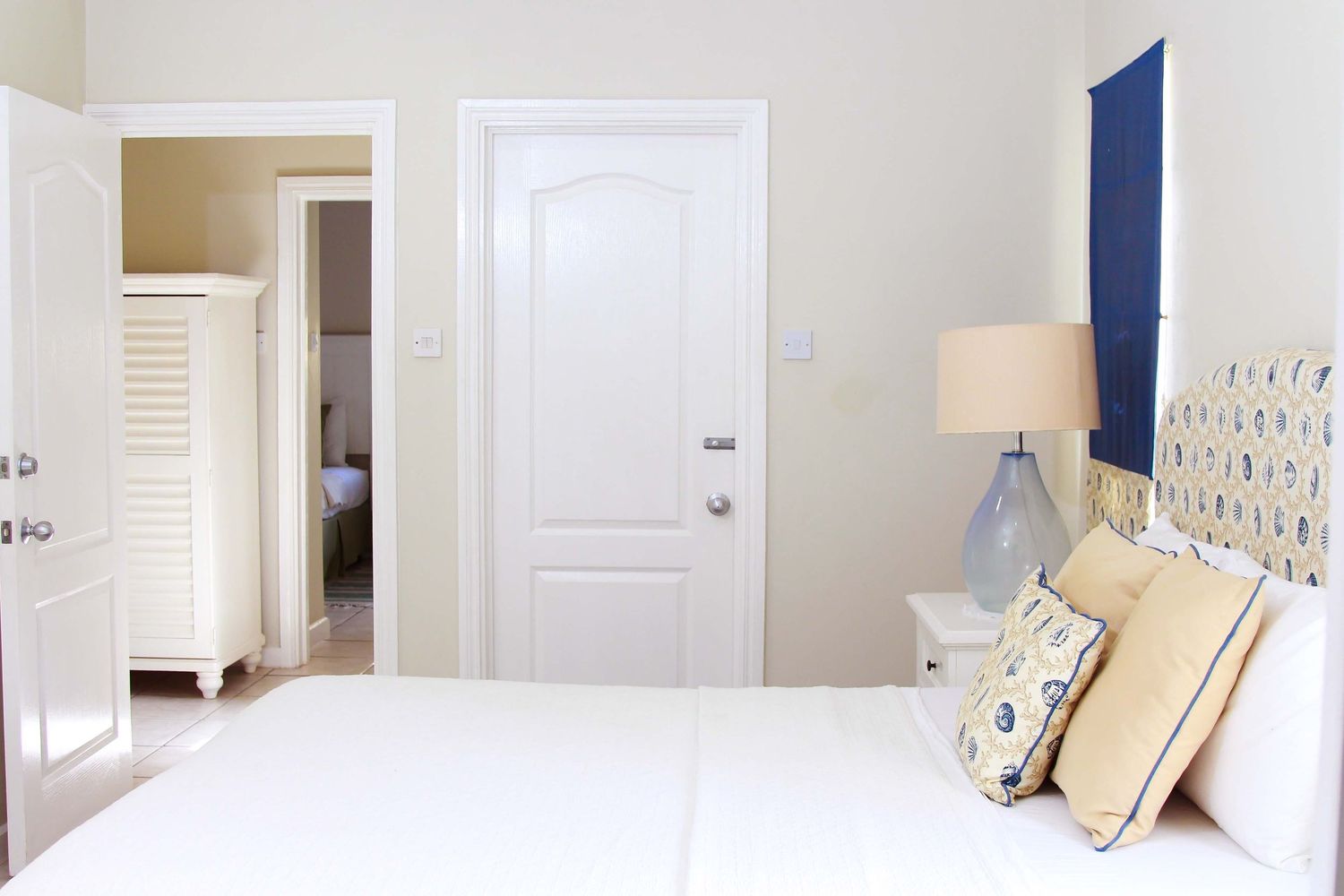 Bright bedroom with white bedding, decorative pillows, a lamp, and a white door leading to a hallway.