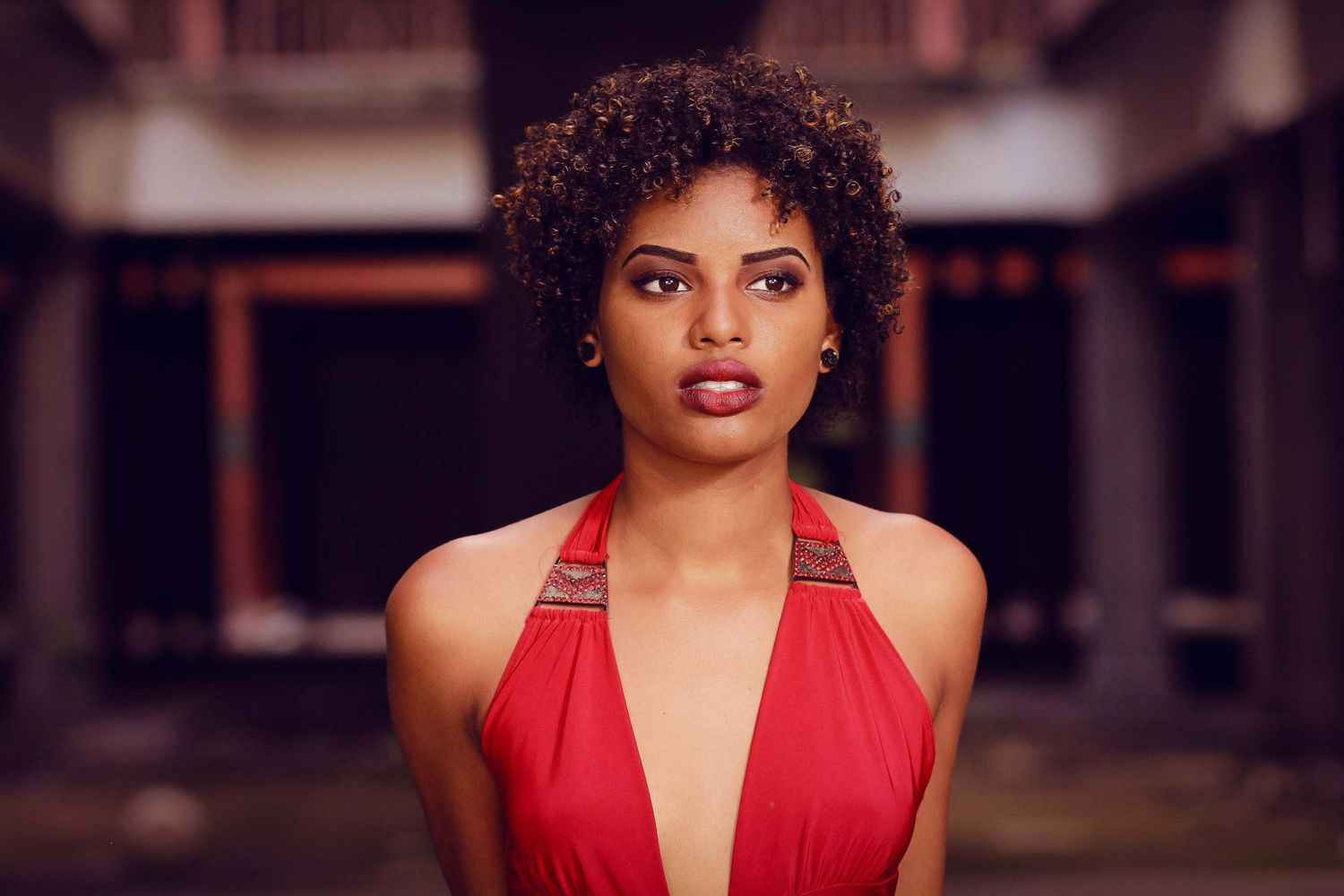 Young woman with curly hair wearing a red halter dress, posing confidently in a dimly lit outdoor setting.