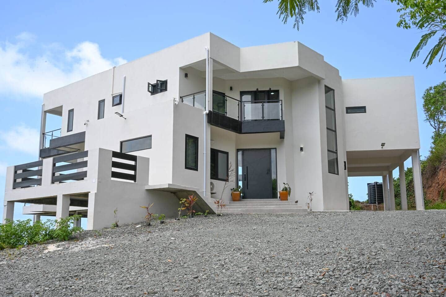 Modern two-story white stucco home with balconies, large windows, and gravel landscaping under a blue sky.