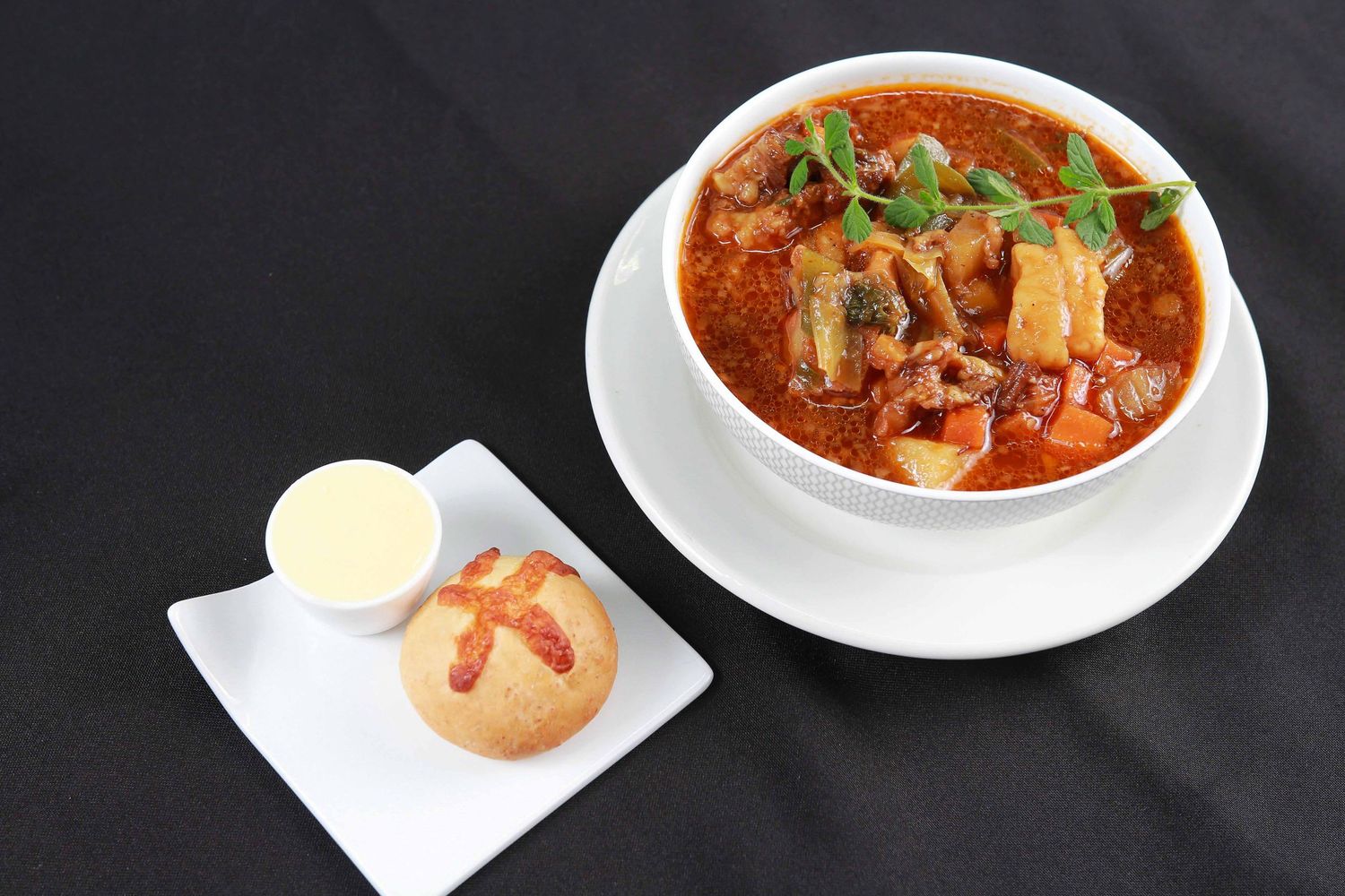 A hearty tomato-based stew with meat and vegetables in a white bowl, served with a dinner roll and dipping sauce.