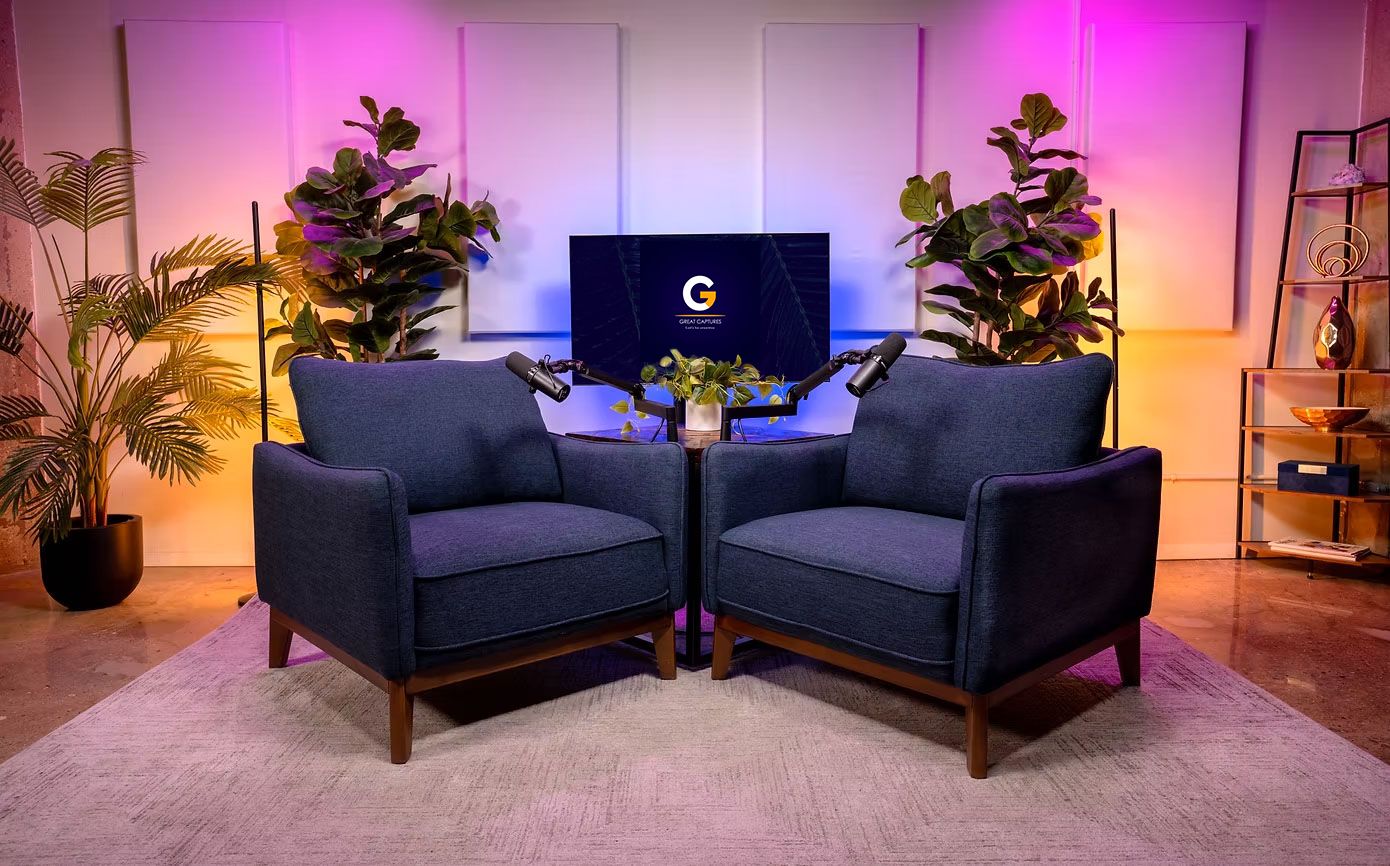 A stylish interview set with two navy blue armchairs, warm purple and yellow lighting, plants, and a screen displaying a logo.