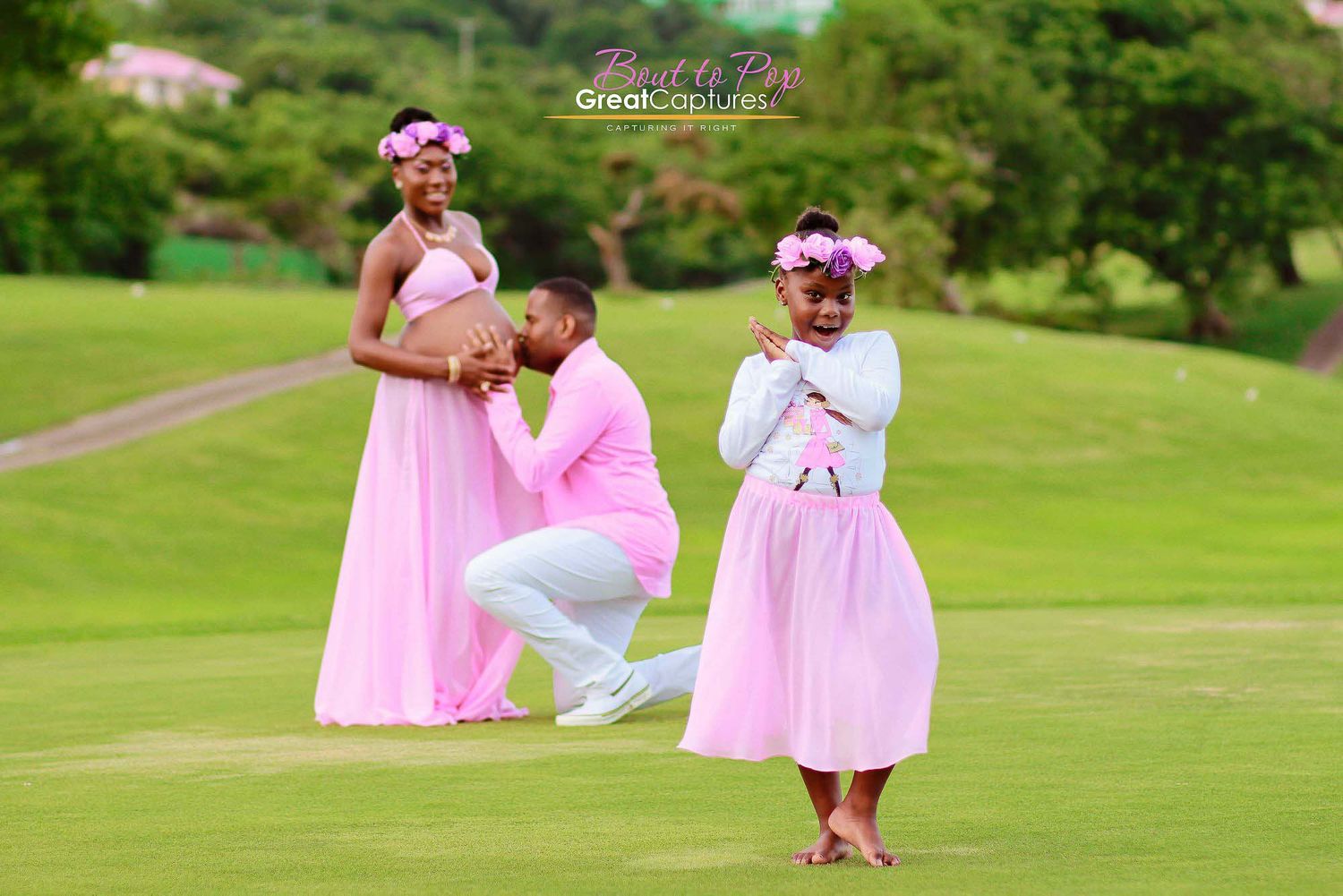 Family maternity photo shoot with couple and child dressed in matching pink outfits on a lush green golf course.