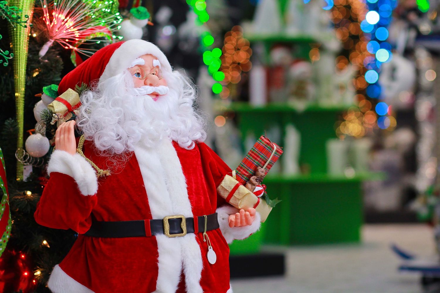 A festive Santa Claus figurine holding gifts stands near a decorated Christmas tree in a colorful holiday display.