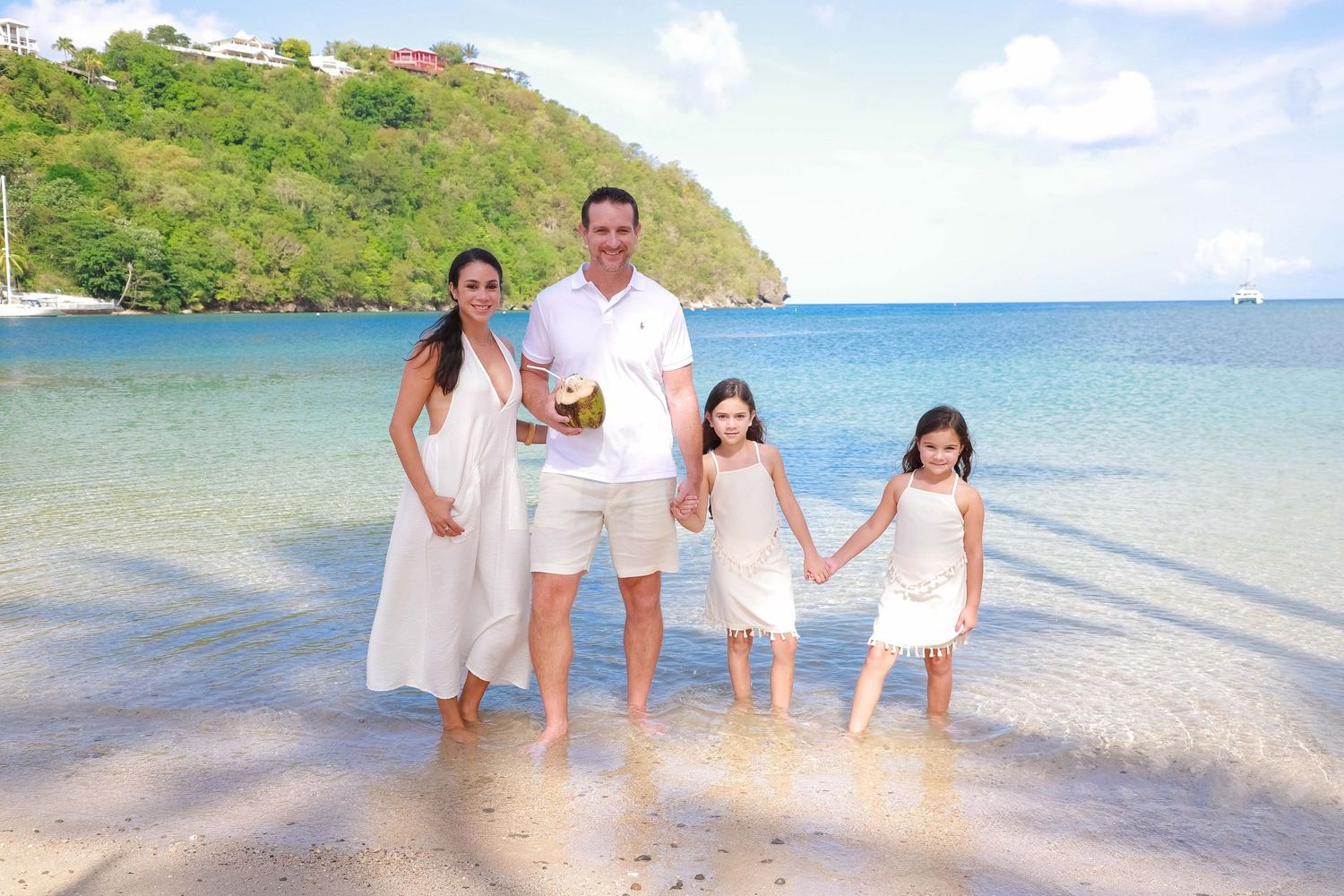 Family of four in white outfits holding hands on a tropical beach with turquoise water and lush green hills.