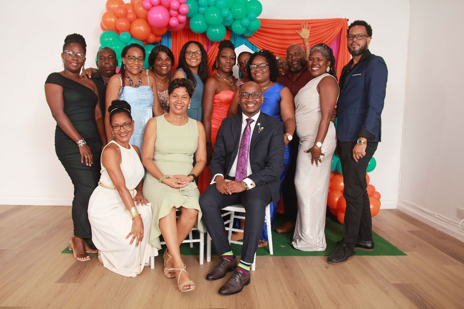 Group of people posing together at a colorful celebration event with balloon decorations in green, orange, and red.