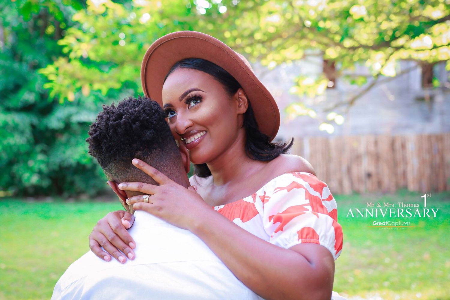 A smiling woman in a floral dress and brown hat embraces a man outdoors during their 1st anniversary celebration.
