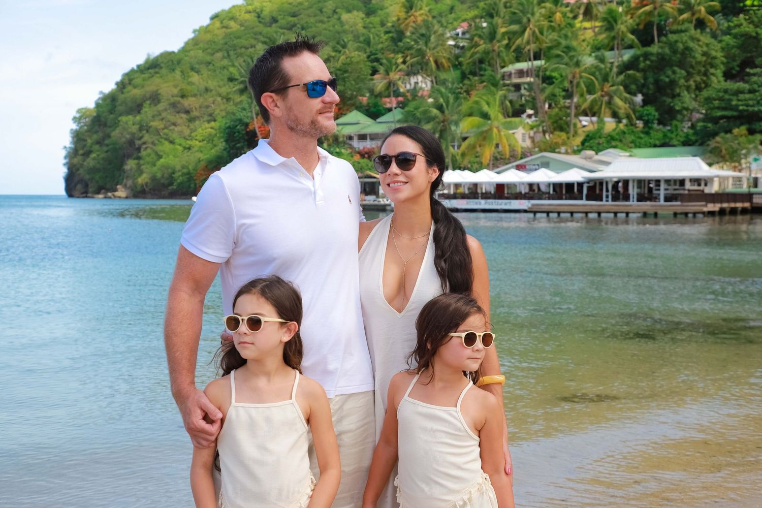 Family of four posing on tropical beach with lush green hills and waterfront resort in background.