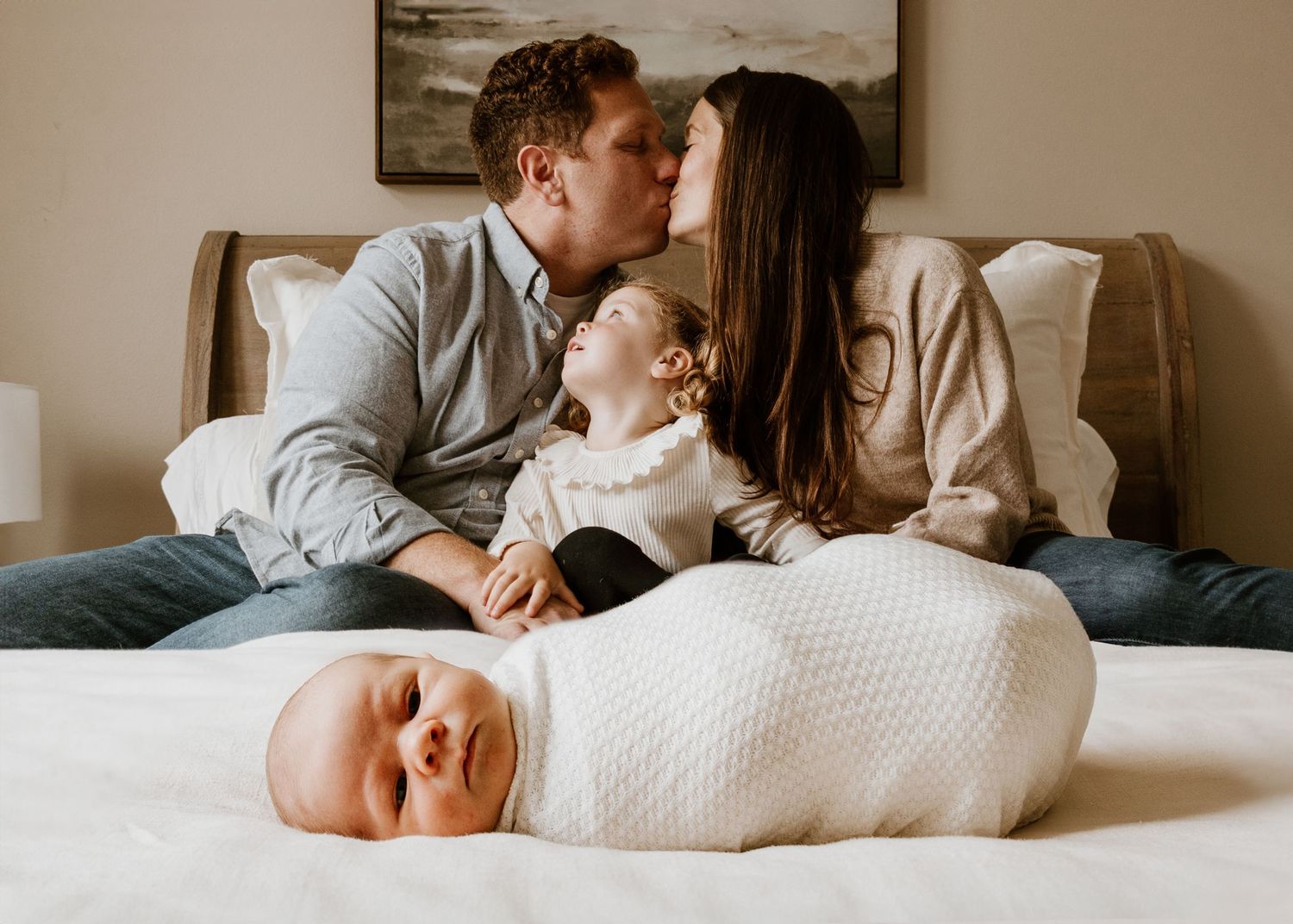 Two parents share a tender moment on a bed as their swaddled baby sleeps peacefully in the foreground.