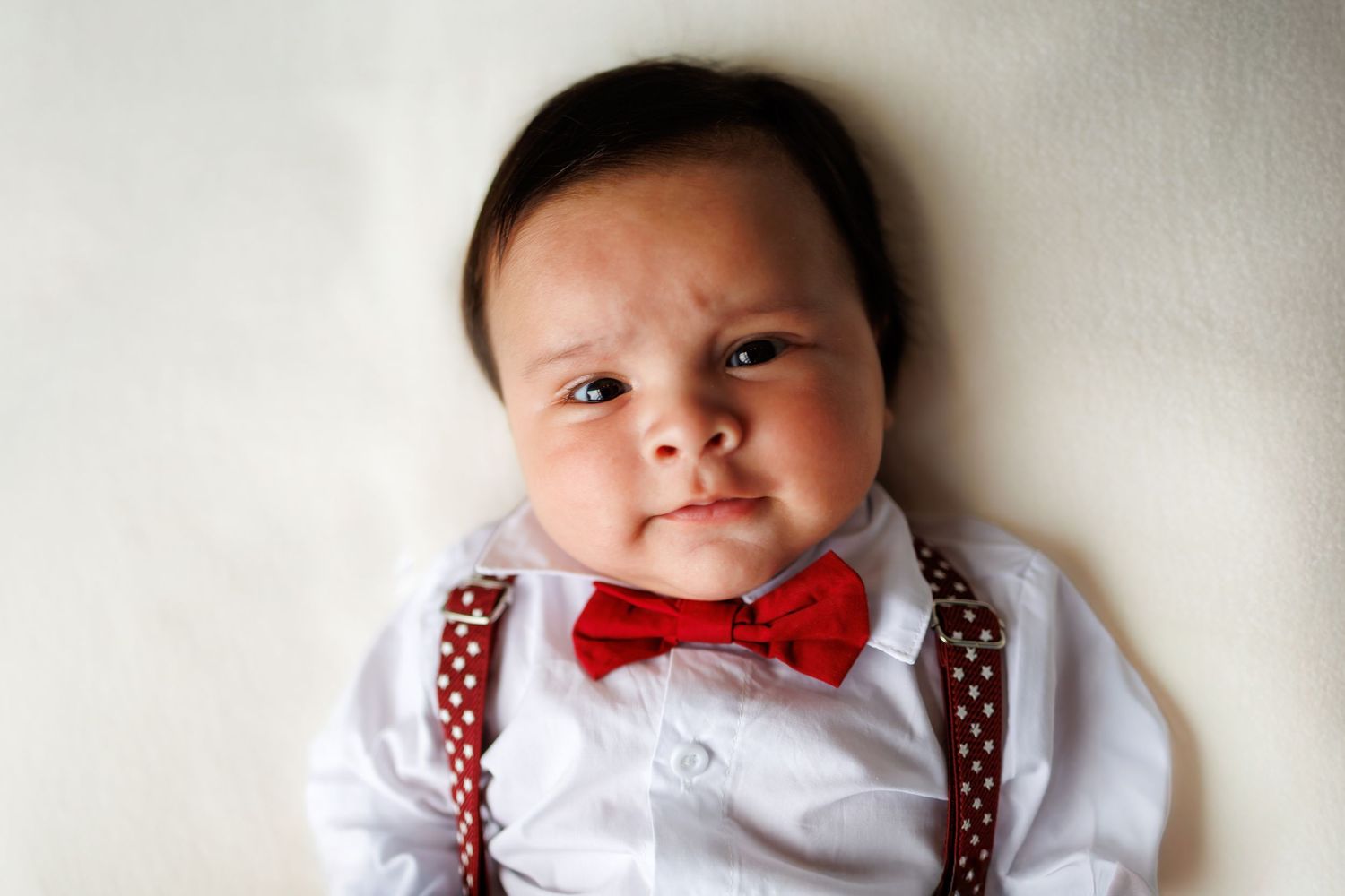 Baby wearing white shirt and red bowtie with brown suspenders poses for three different profile shots.