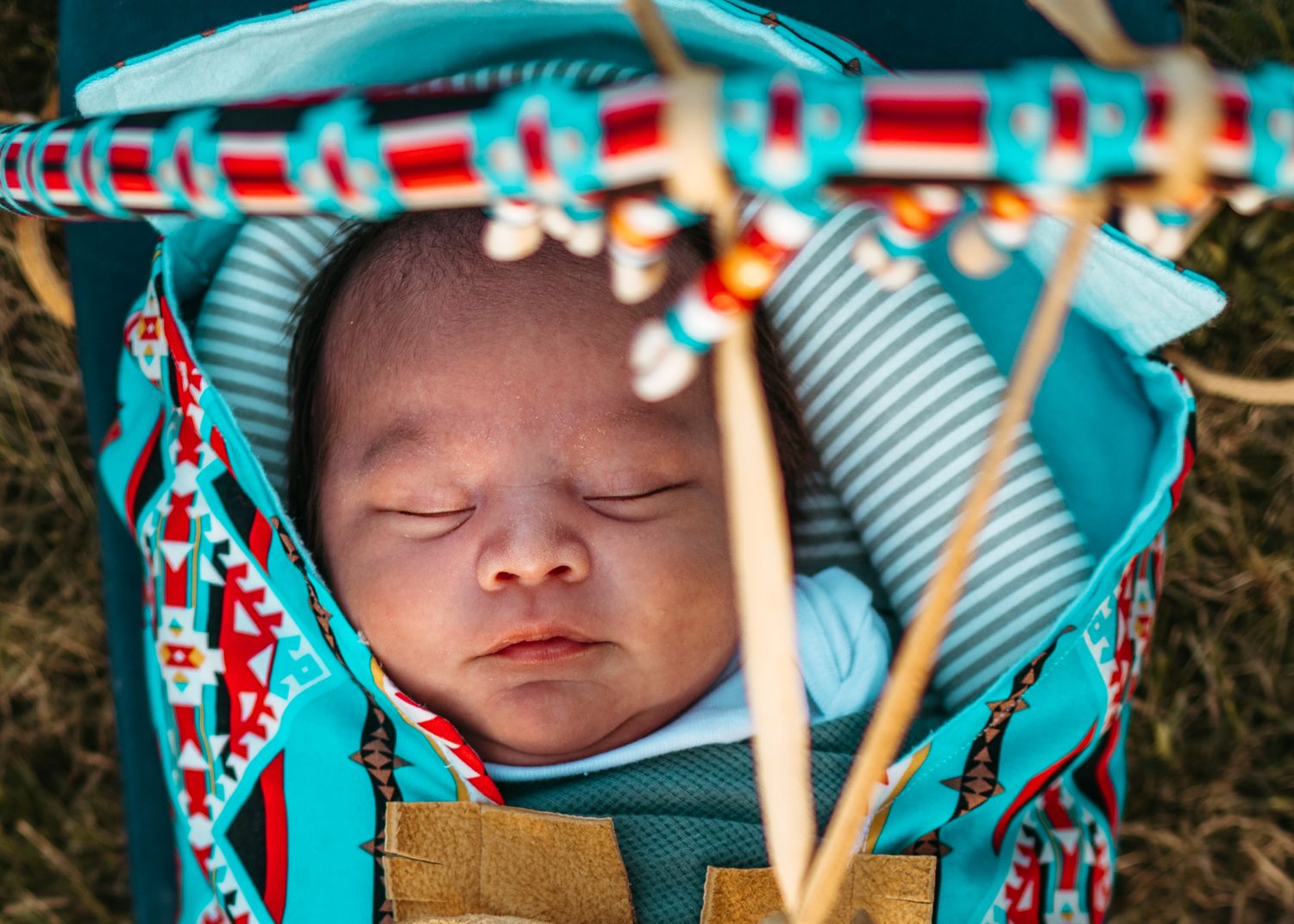 Teal and red patterned baby carrier holding sleeping infant outdoors with natural background.