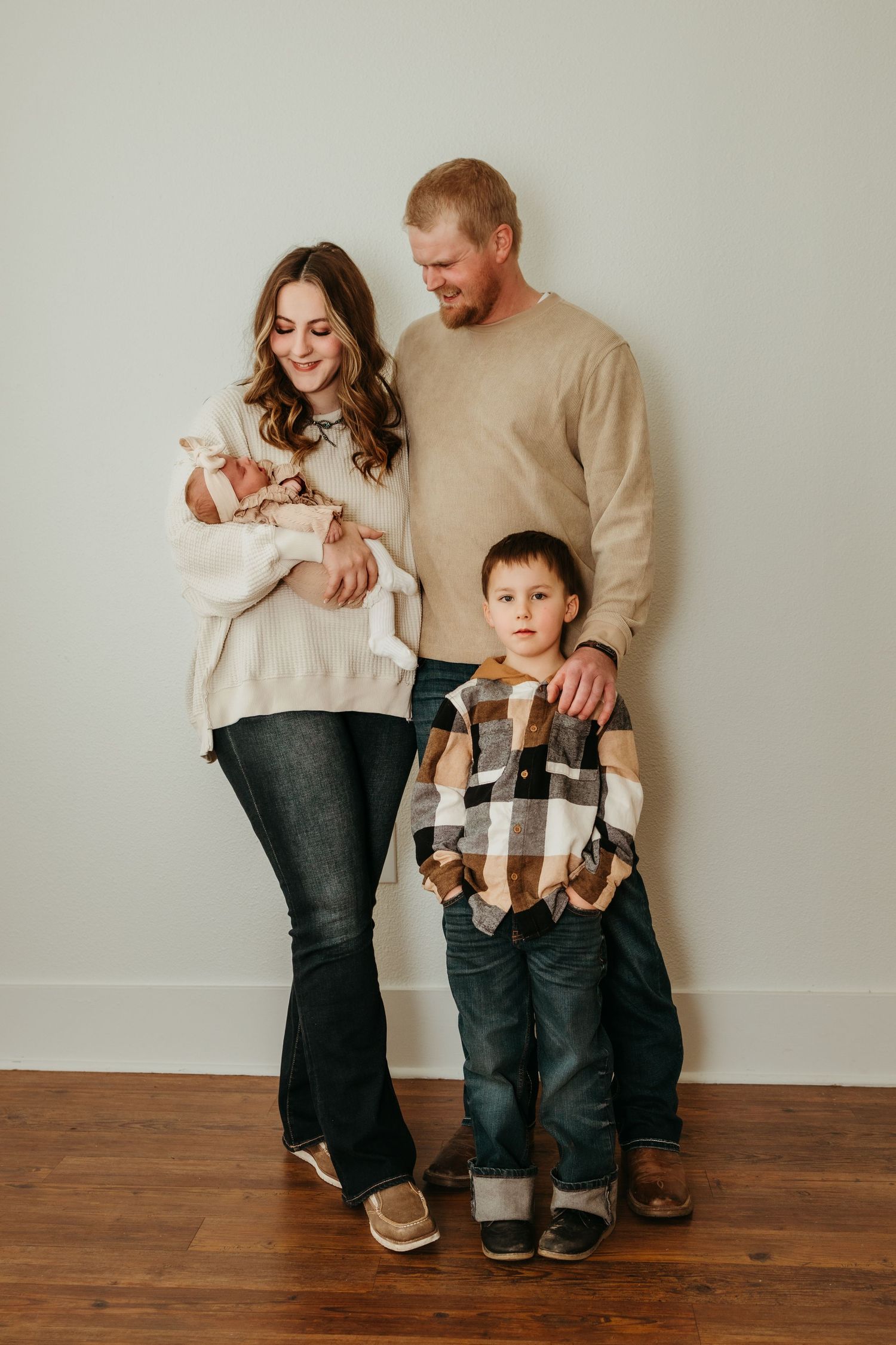 A family of three stands together on hardwood floors wearing coordinated neutral and plaid outfits in a studio setting.