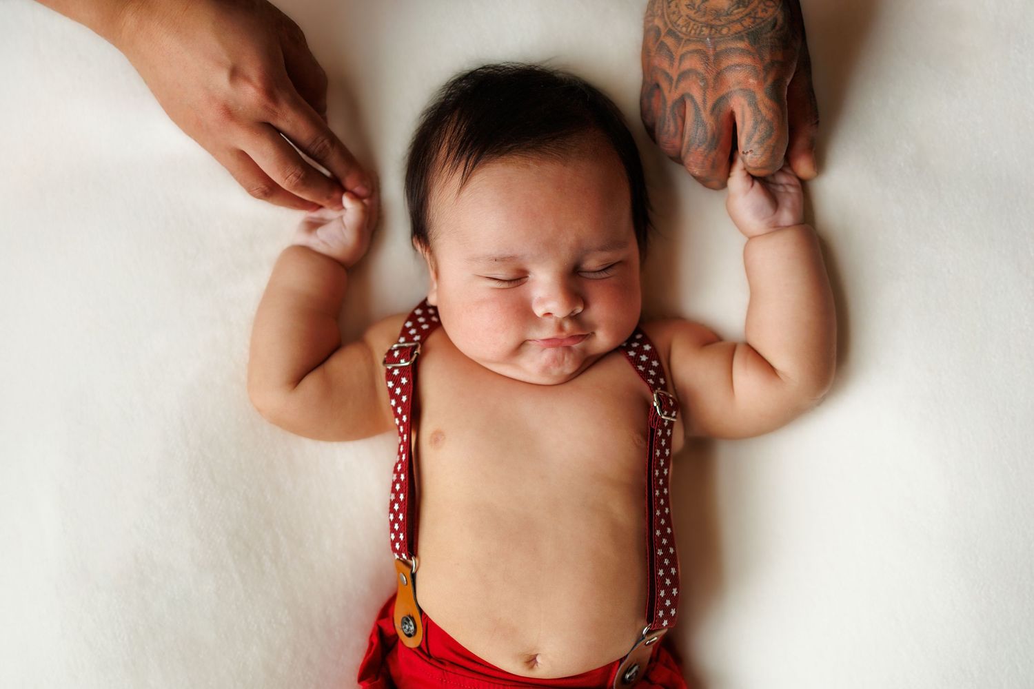 A sleeping baby in red suspenders lies peacefully on white blankets while hands gently touch their arms.