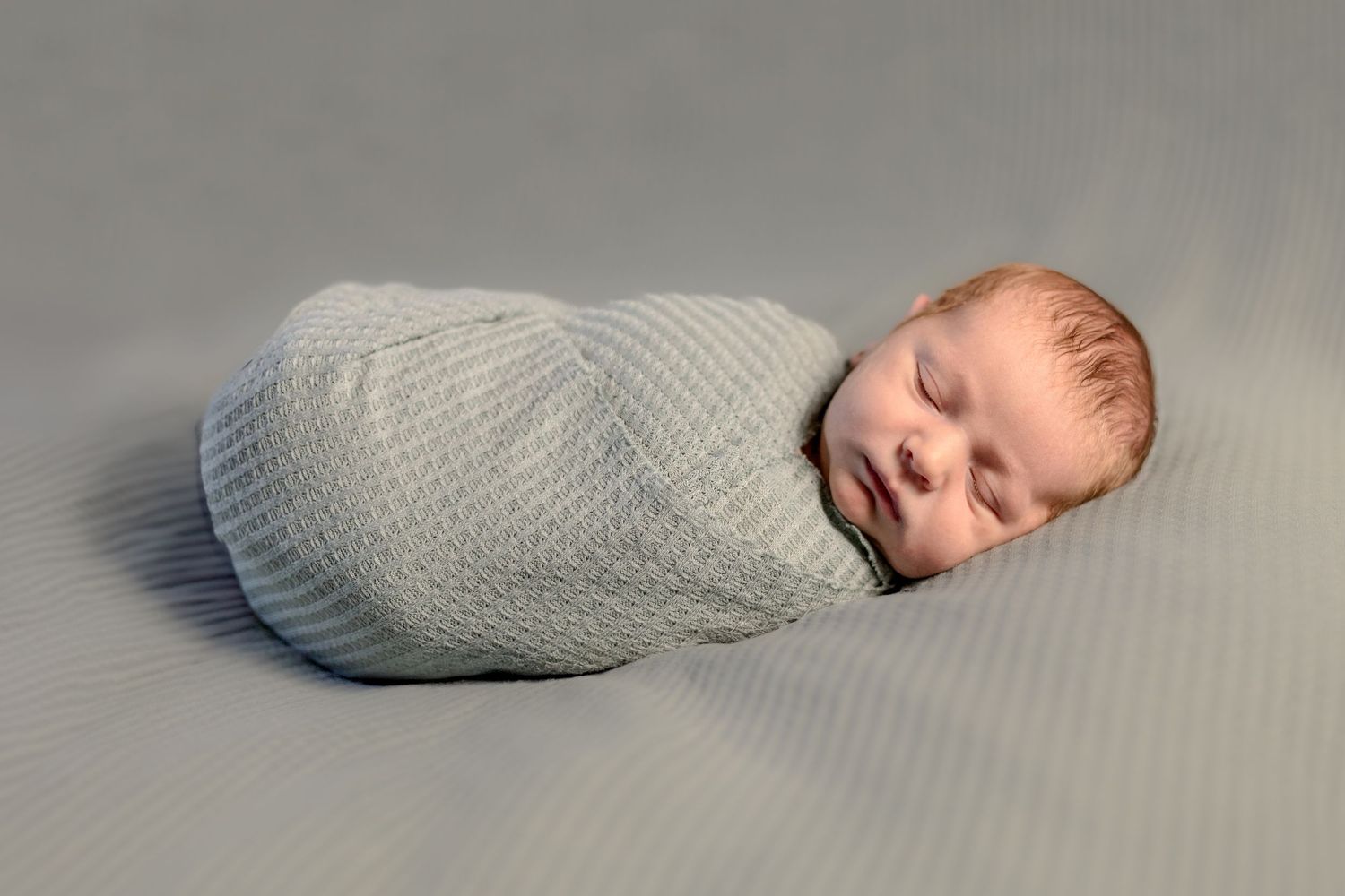 Peaceful sleeping newborn wrapped in soft gray textured swaddle blanket against neutral backdrop.