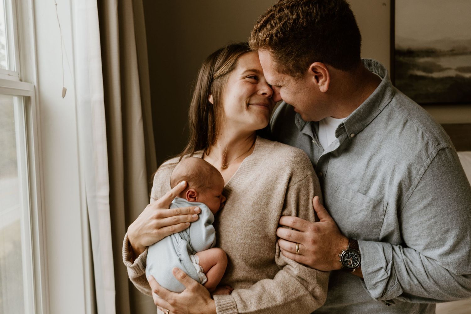 A tender family moment by the window as parents share a loving embrace while holding their sleeping newborn baby.
