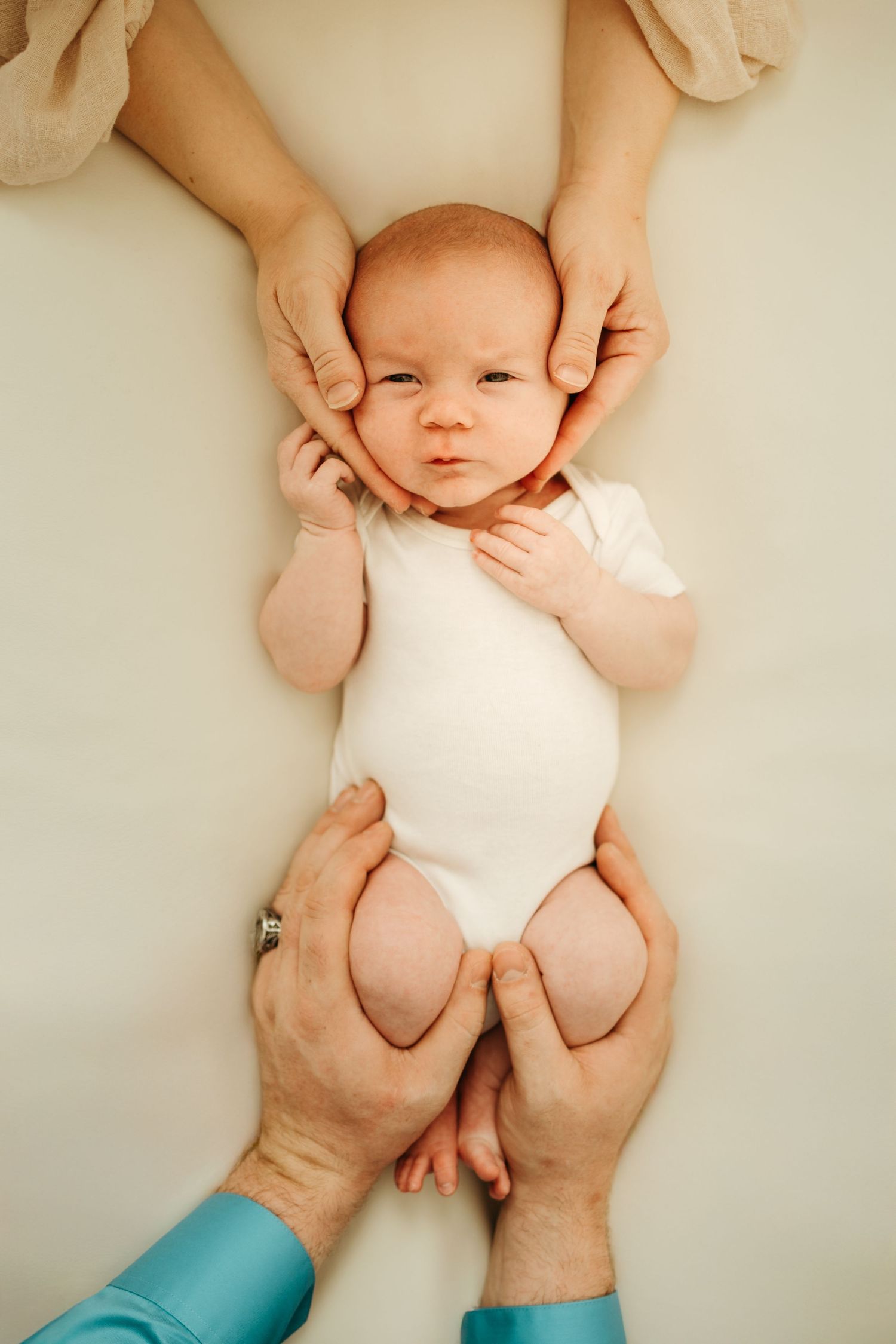 Tender hands hold a sleeping newborn baby swaddled in white fabric against a neutral background.