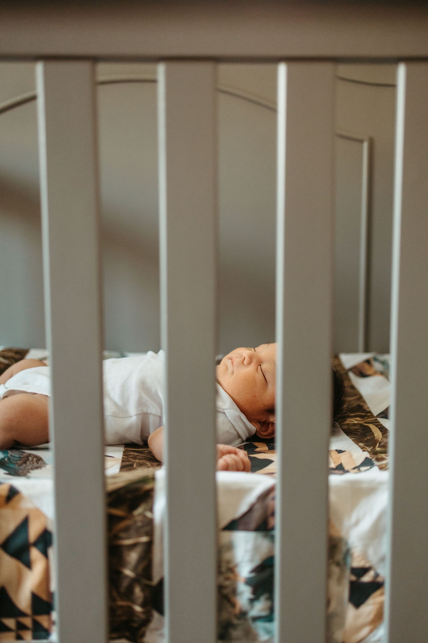 A view through white crib bars showing someone sleeping peacefully under blankets and pillows.