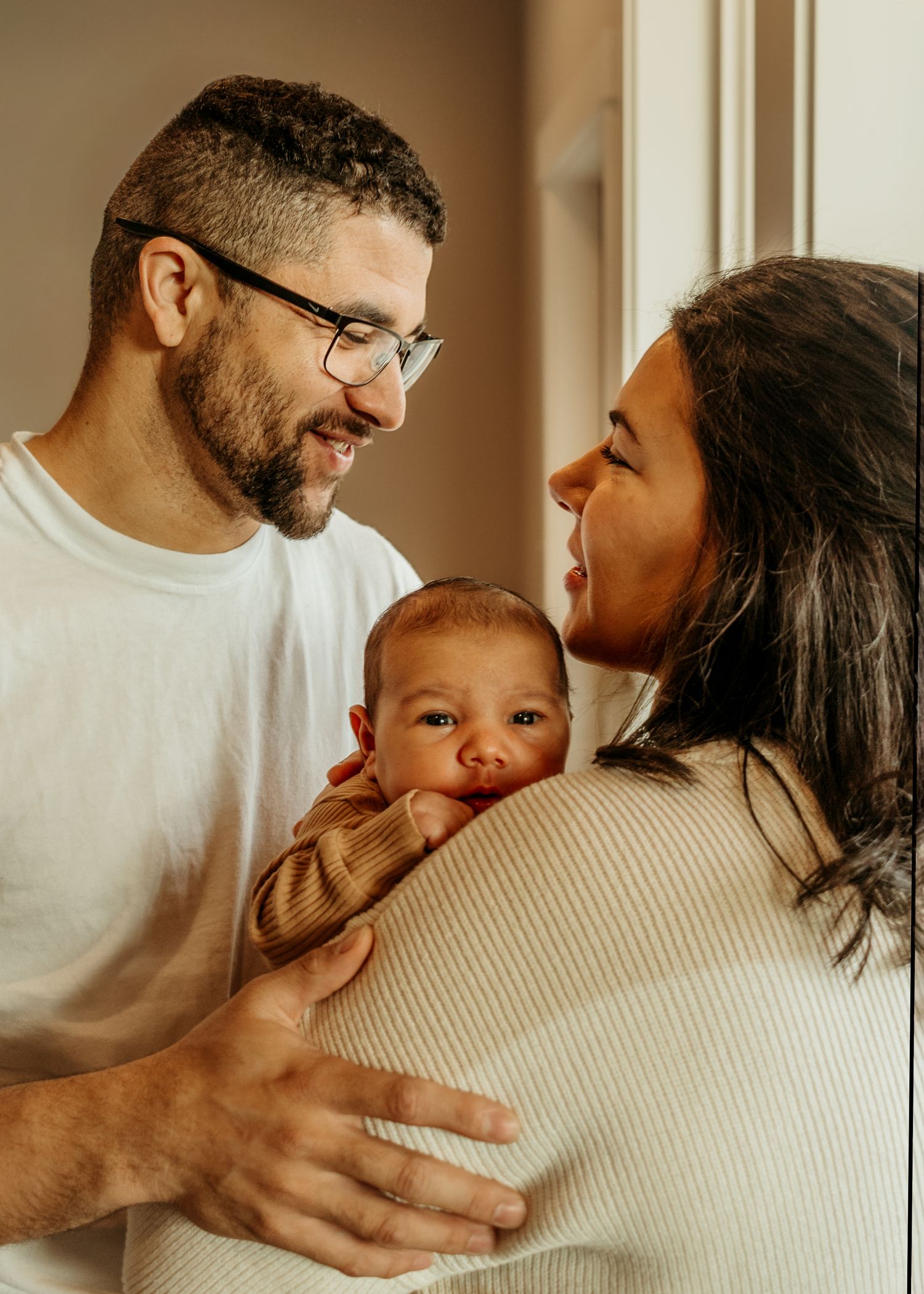 Young family looking lovingly at their adorable baby while standing in warm natural light near a window.