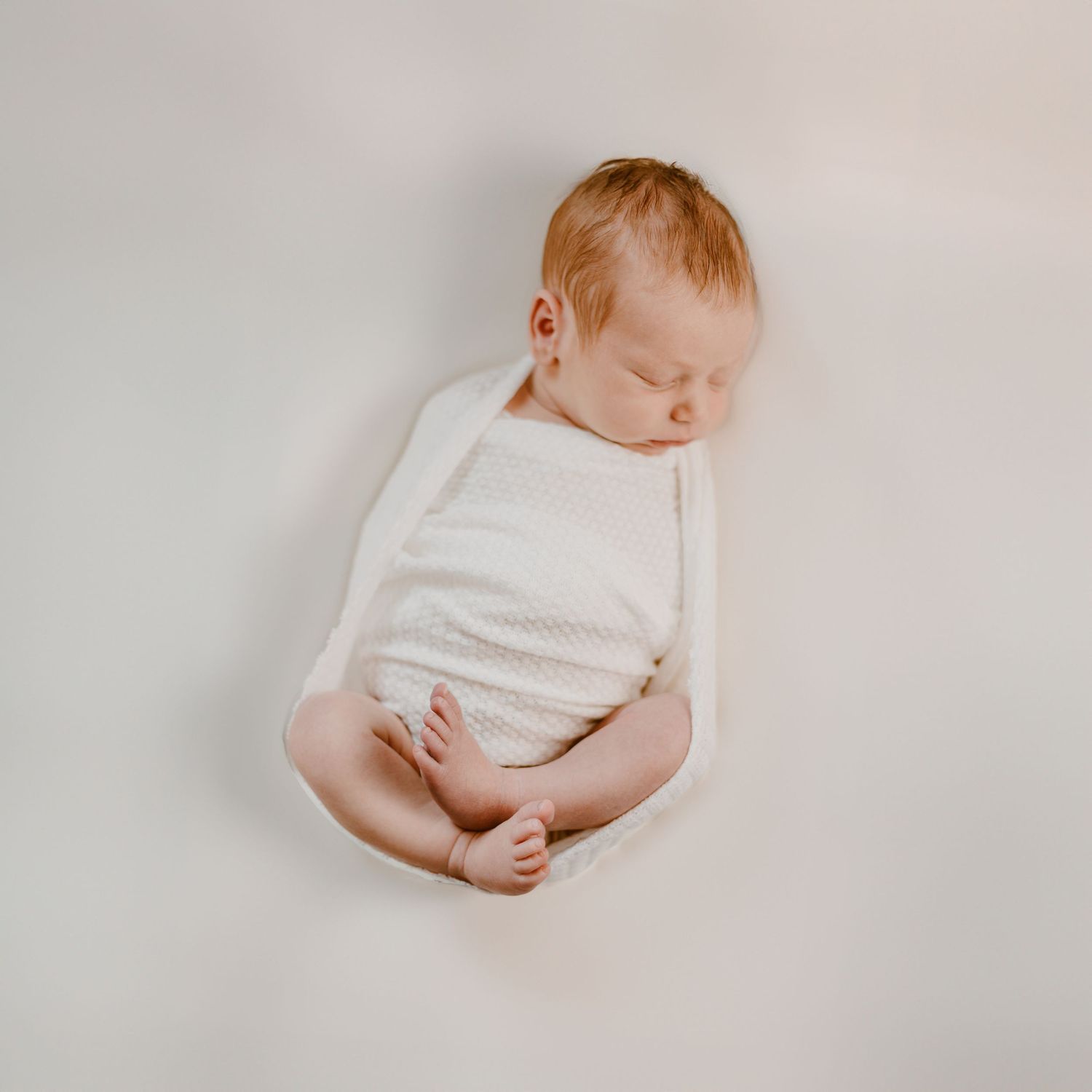 A sleeping newborn baby wearing white clothing lies peacefully against a light neutral background.