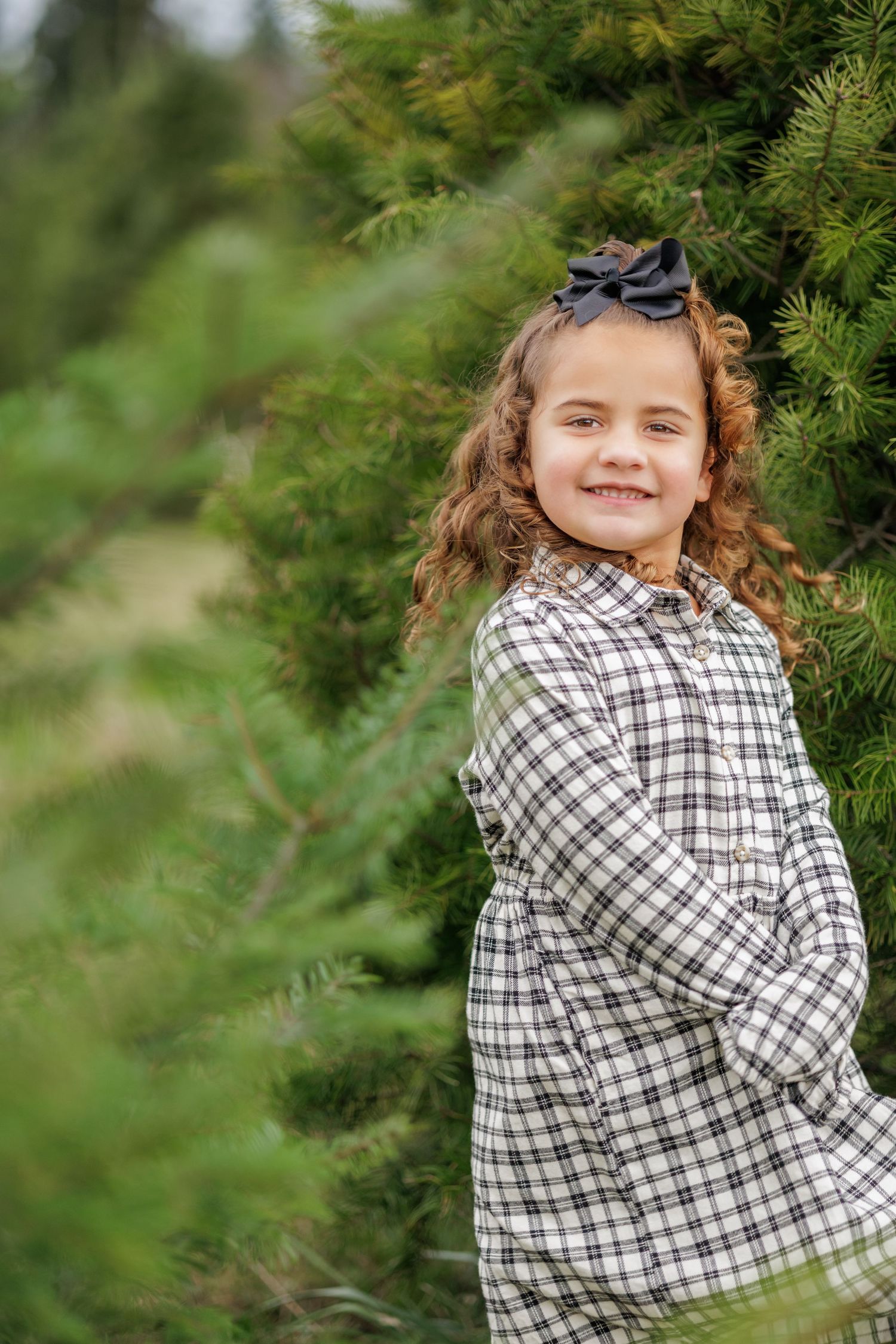 A child wearing a black and white plaid dress poses playfully near evergreen trees in an outdoor setting.