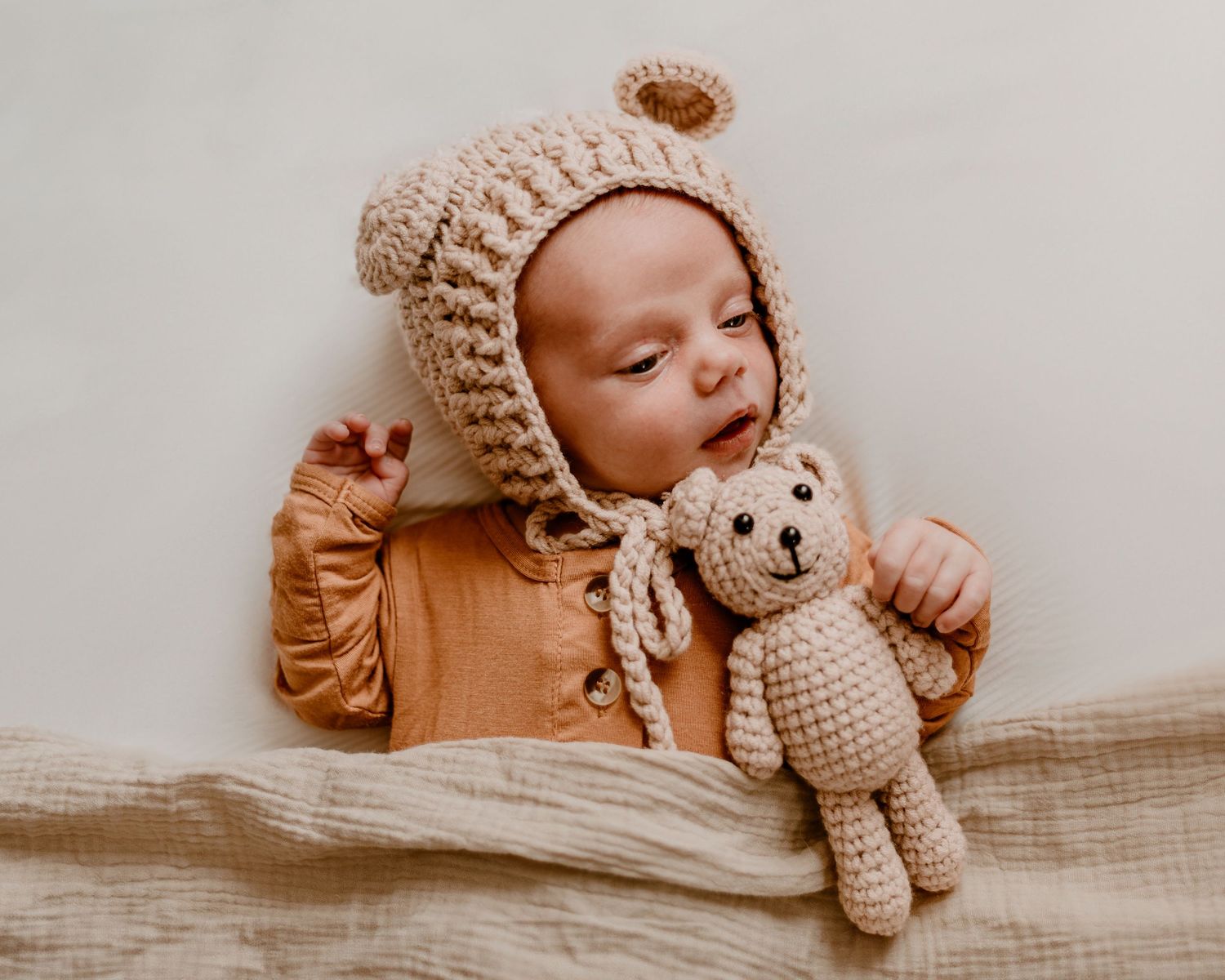 Sweet newborn photography setup featuring a sleeping baby with soft knit bonnet and teddy bear on neutral bedding.