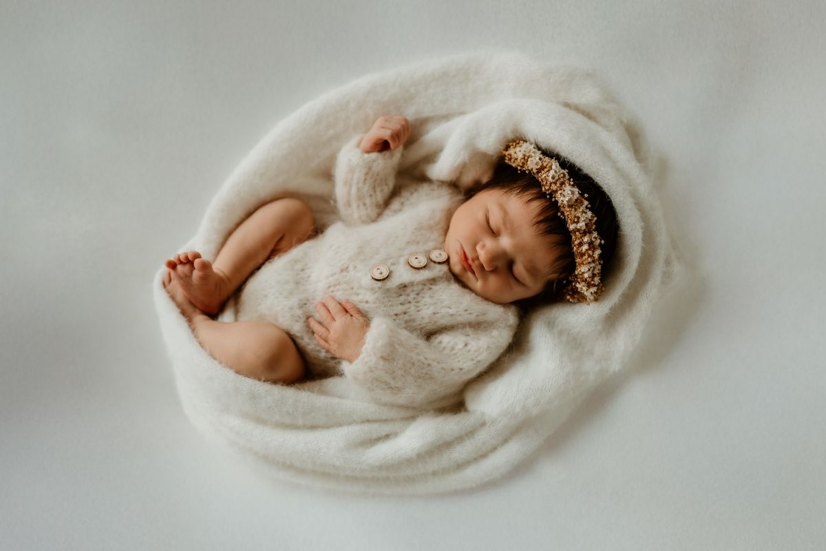 Sleeping newborn baby in cream knit outfit and floral headband, nestled in a soft white blanket.