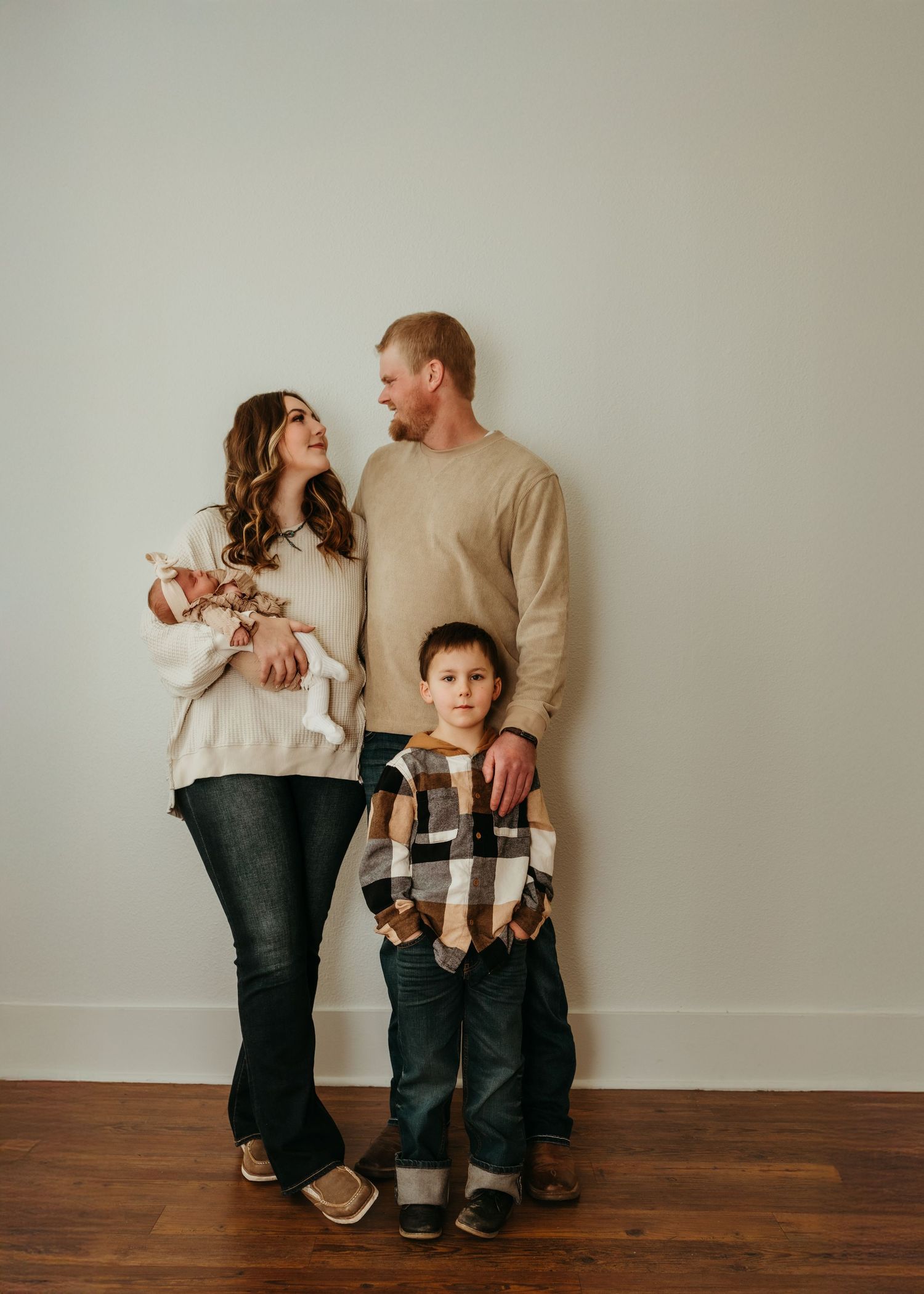People in matching beige sweaters and jeans sit together on a hardwood floor against a white wall.