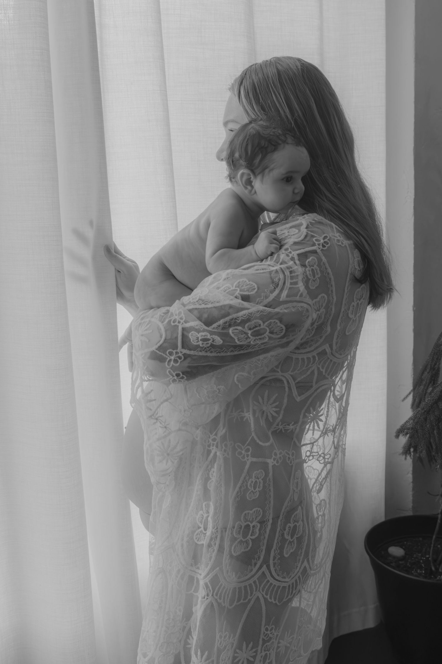 A tender black and white portrait of mother and baby near a curtained window, with the mother wearing a detailed lace dress.