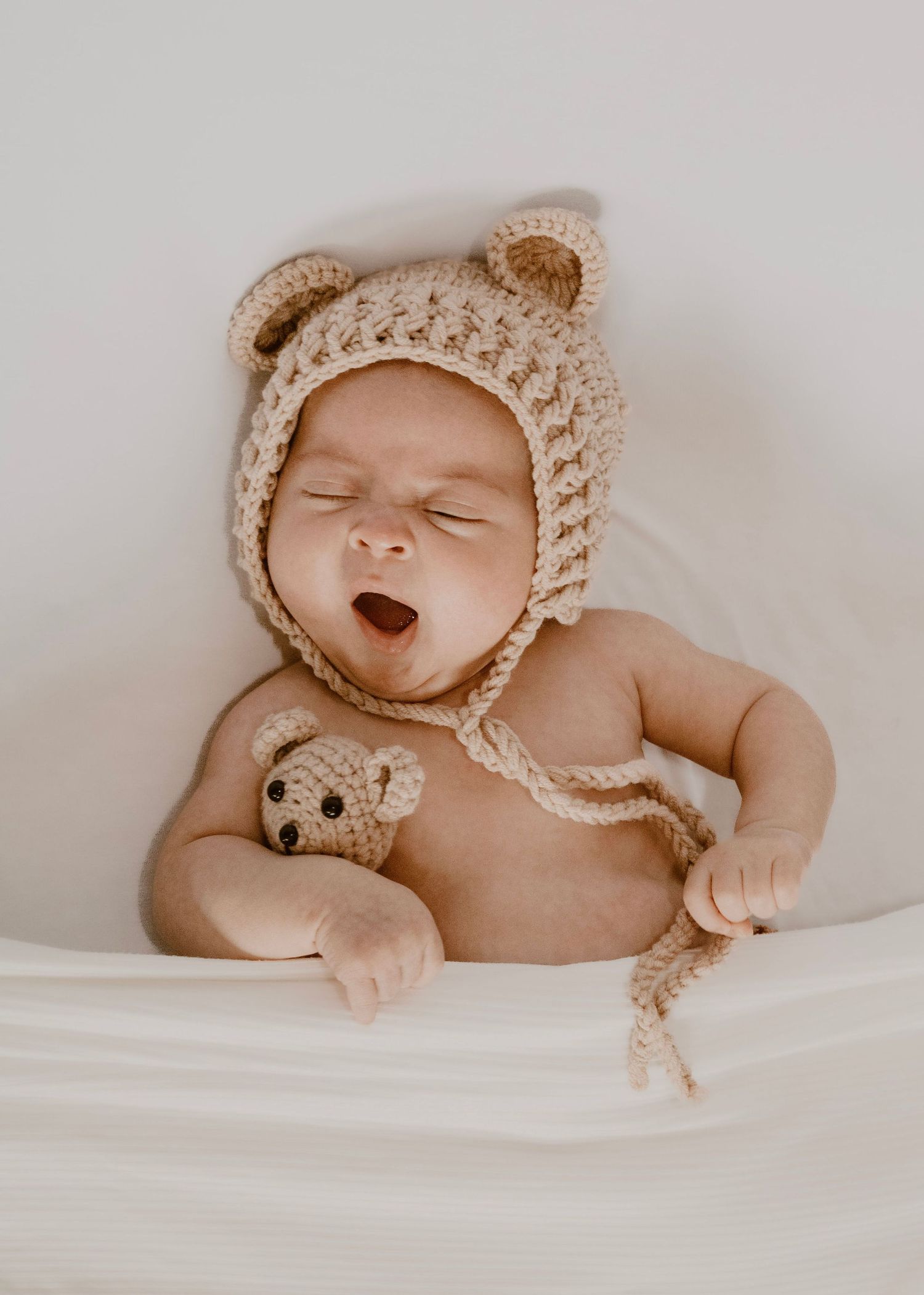 Sleeping baby wearing a knitted beige bear bonnet lies on white bedding while holding a small teddy bear toy.
