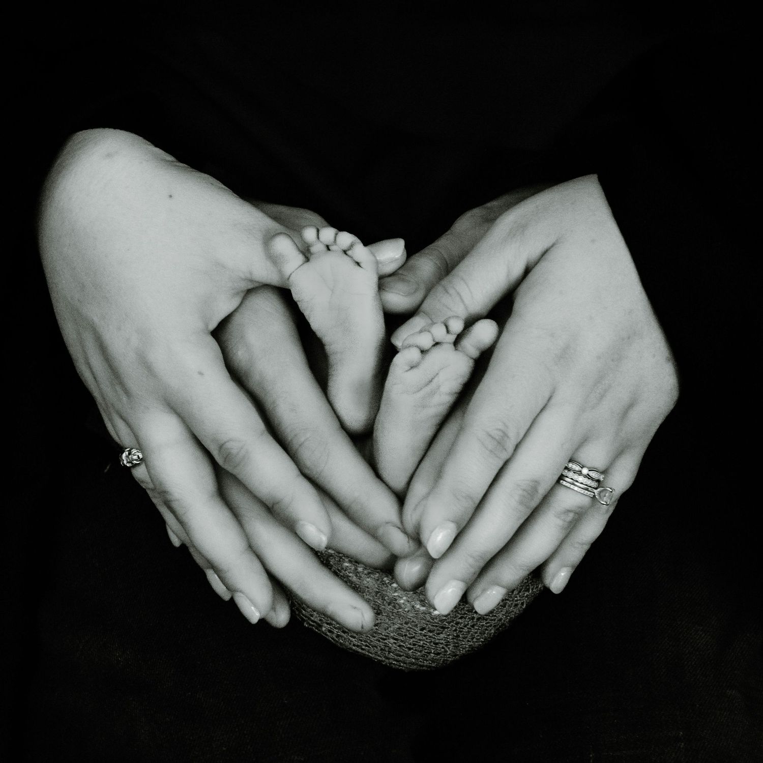Black and white close-up photo of tender adult hands cradling tiny baby feet with wedding rings visible.