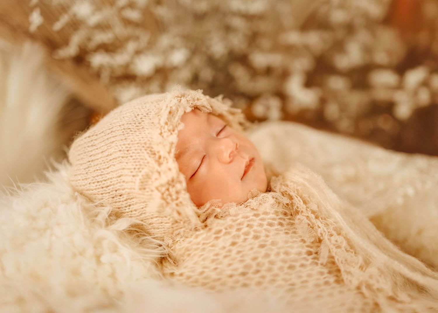 A sleeping baby swaddled in cream knitted blankets and wearing a matching knit bonnet lies peacefully on soft fabric.
