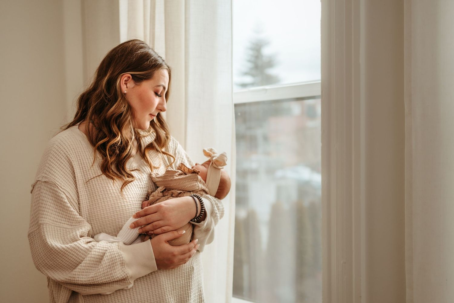 Woman dressed in cozy cream sweater holds sleeping newborn baby in serene home setting by window.