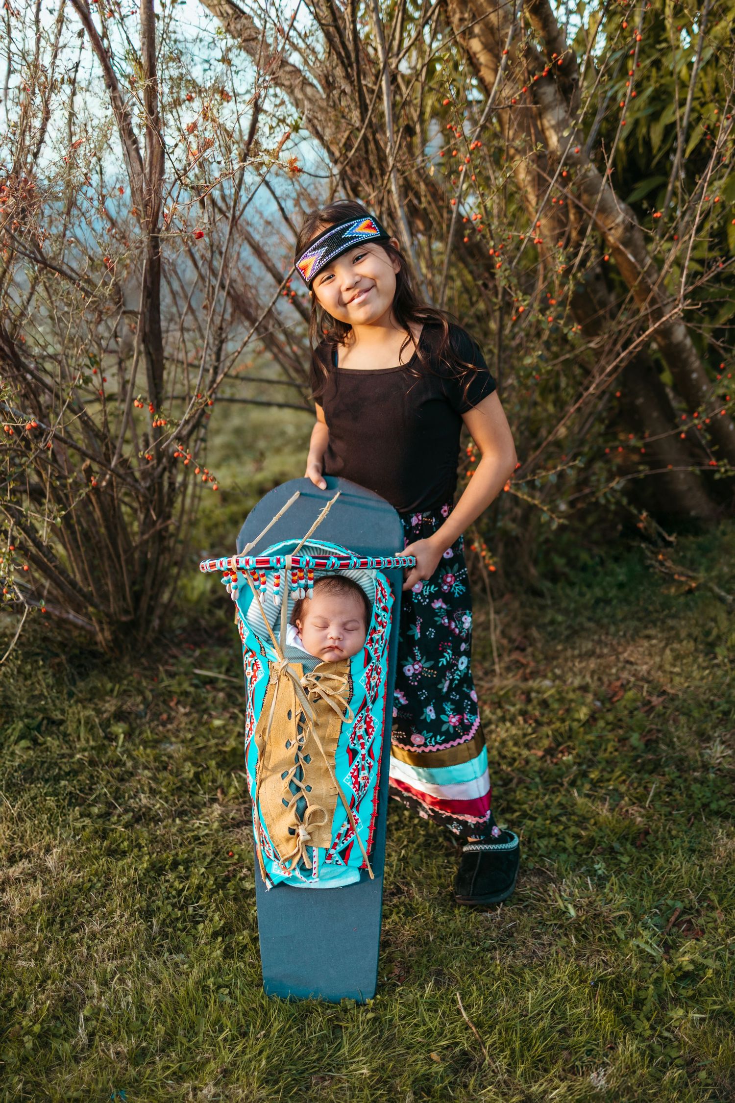 Two people in traditional Native American regalia pose together outdoors with decorative blue cradleboard against natural backdrop.