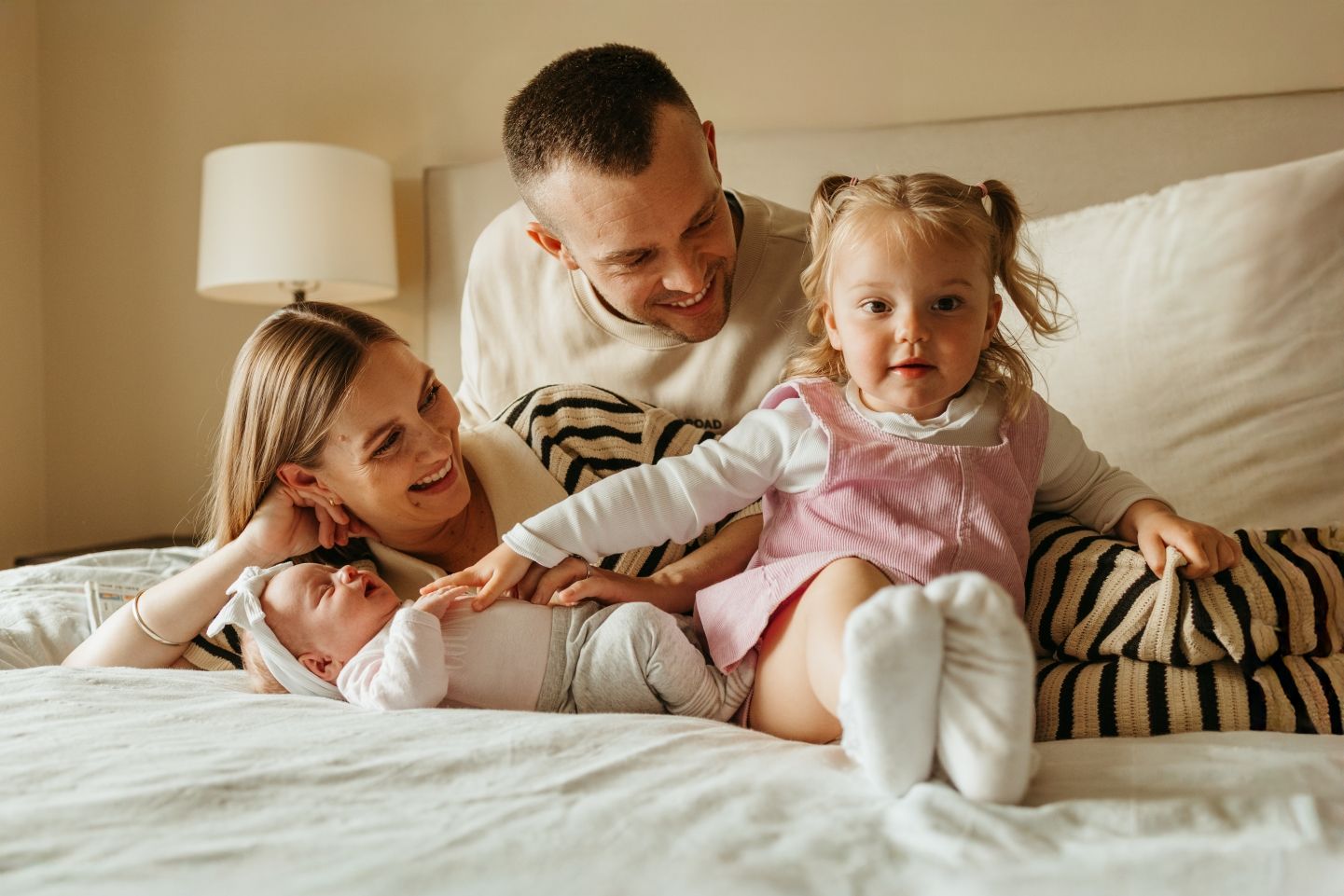 Happy family of four relaxing on a bed, parents smiling at newborn baby while toddler girl sits nearby.