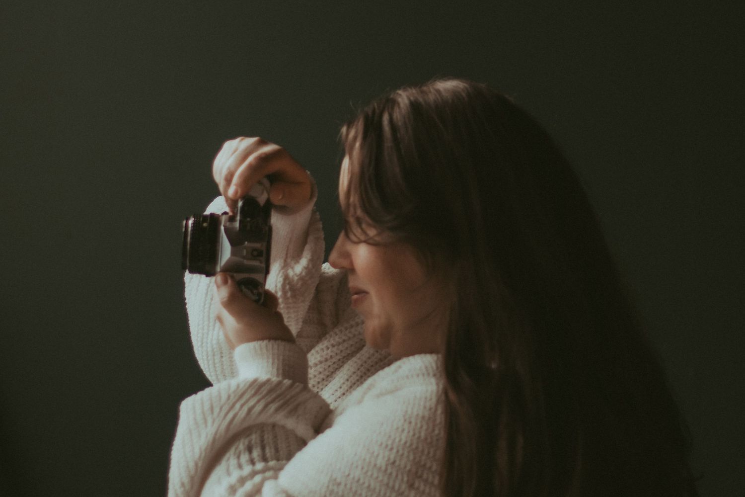 A photographer wearing a cream sweater holds up a vintage camera in moody lighting.