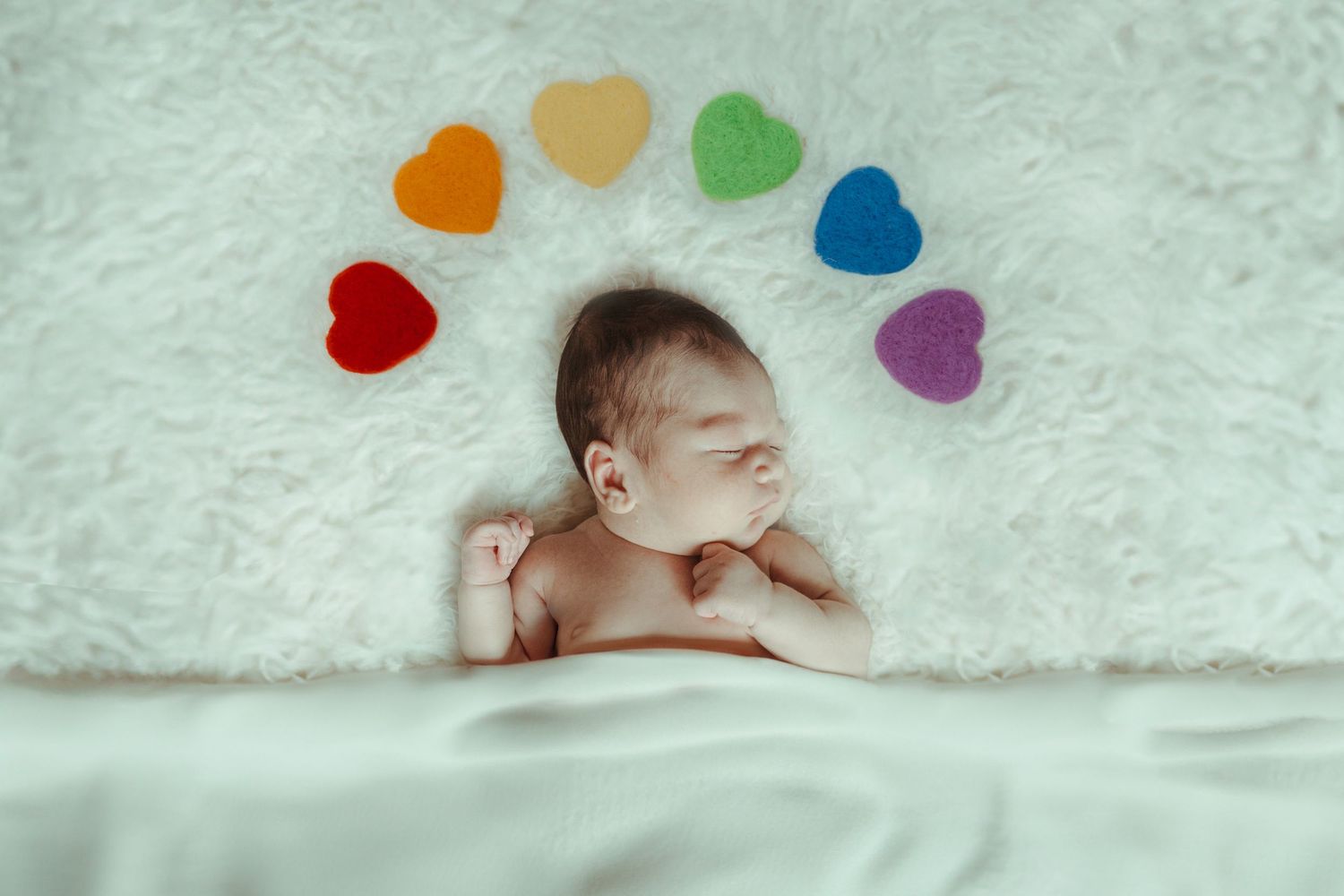Sleeping baby photographed on white blanket with colorful rainbow heart shapes arranged in an arc above.