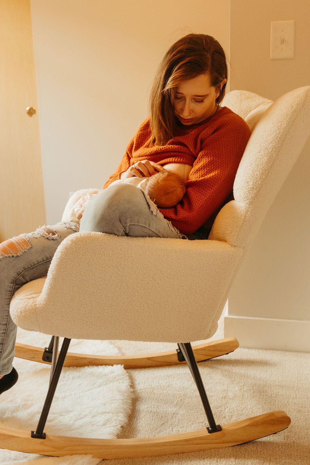 Person in an orange sweater sits in a modern white rocking chair while nursing a baby in a cozy home setting.