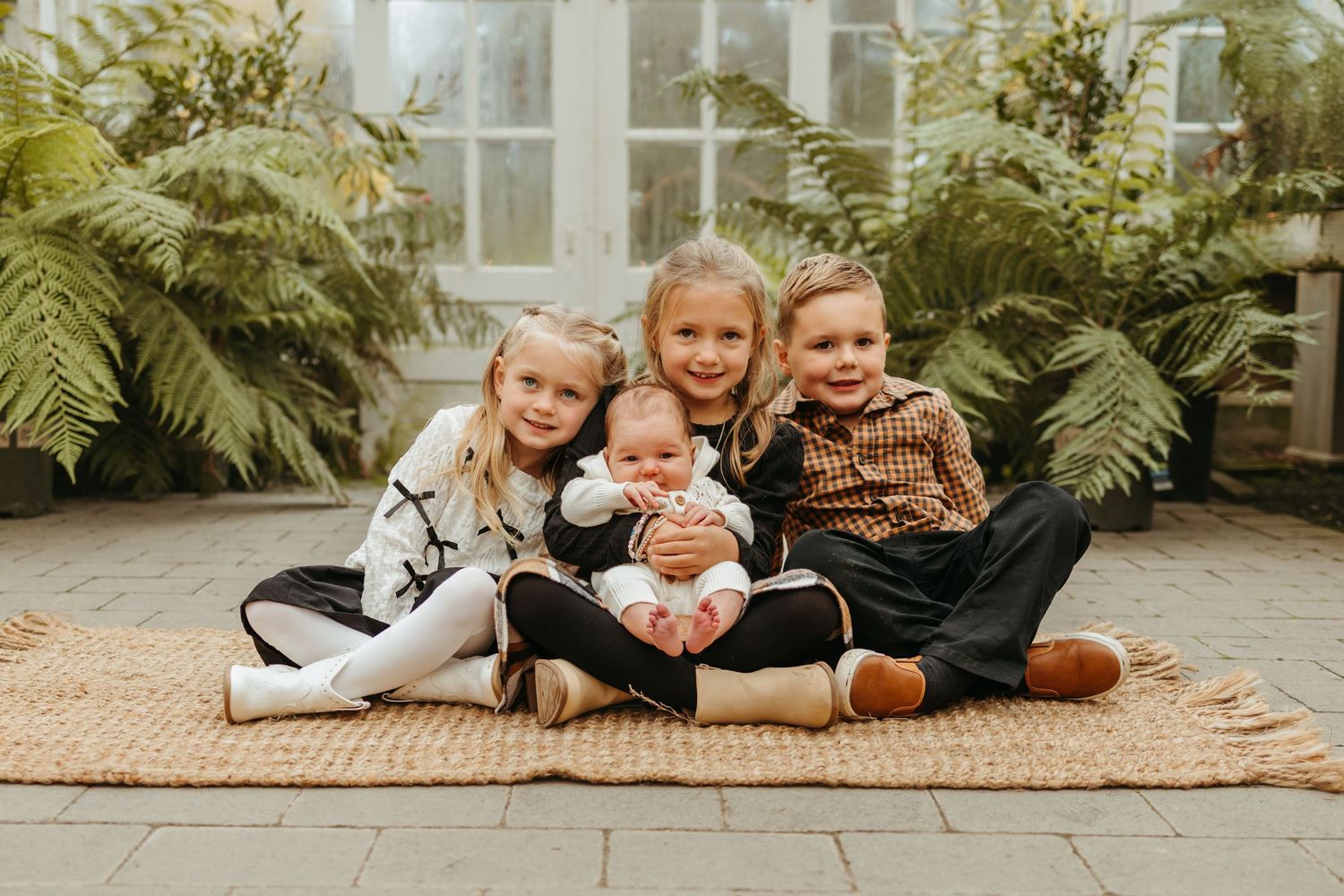 Children laying on a woven rug in a greenhouse setting with lush ferns and plants in the background.
