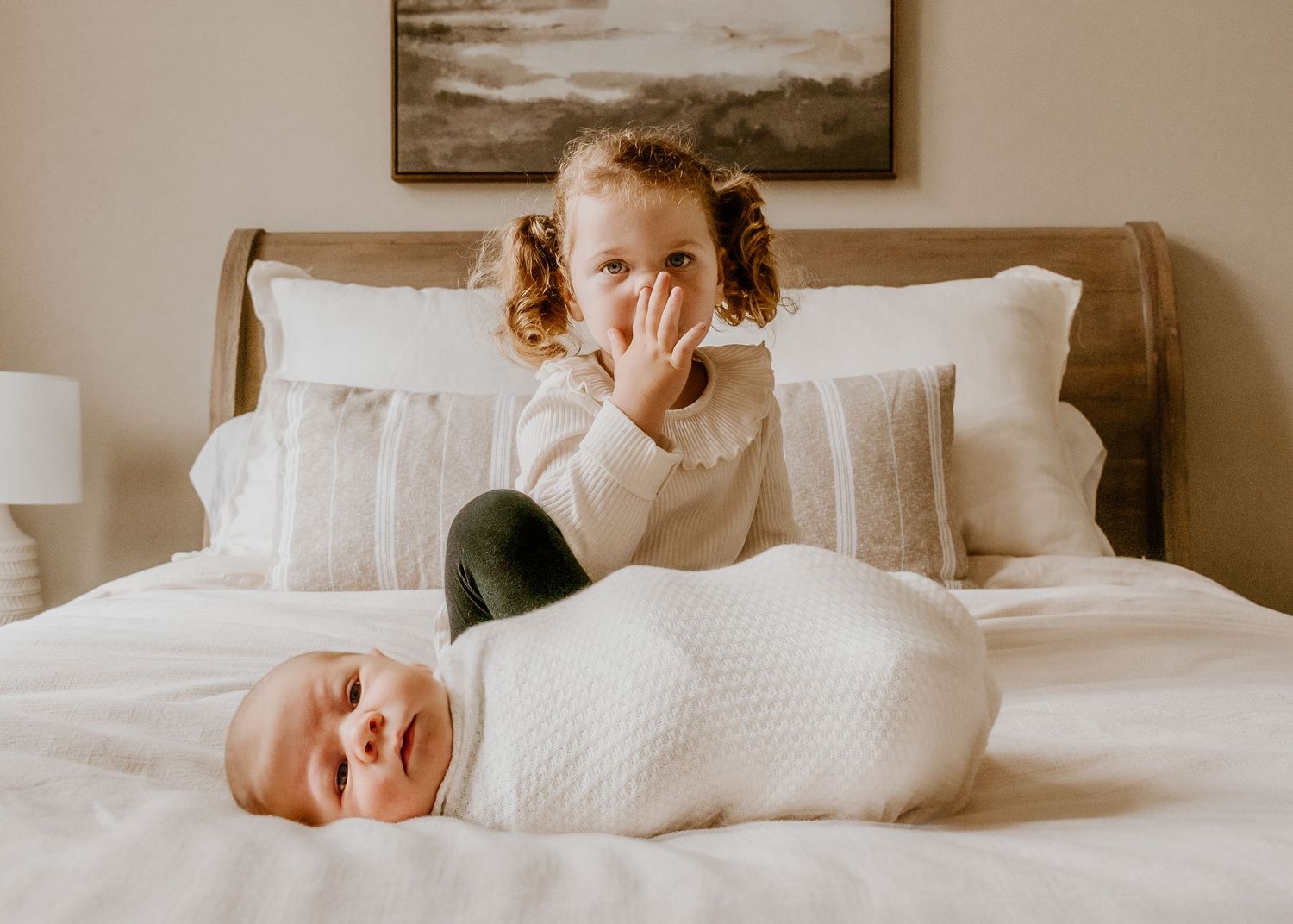 Two siblings on a white bed with pillows sitting together in a cozy bedroom with neutral beige walls.